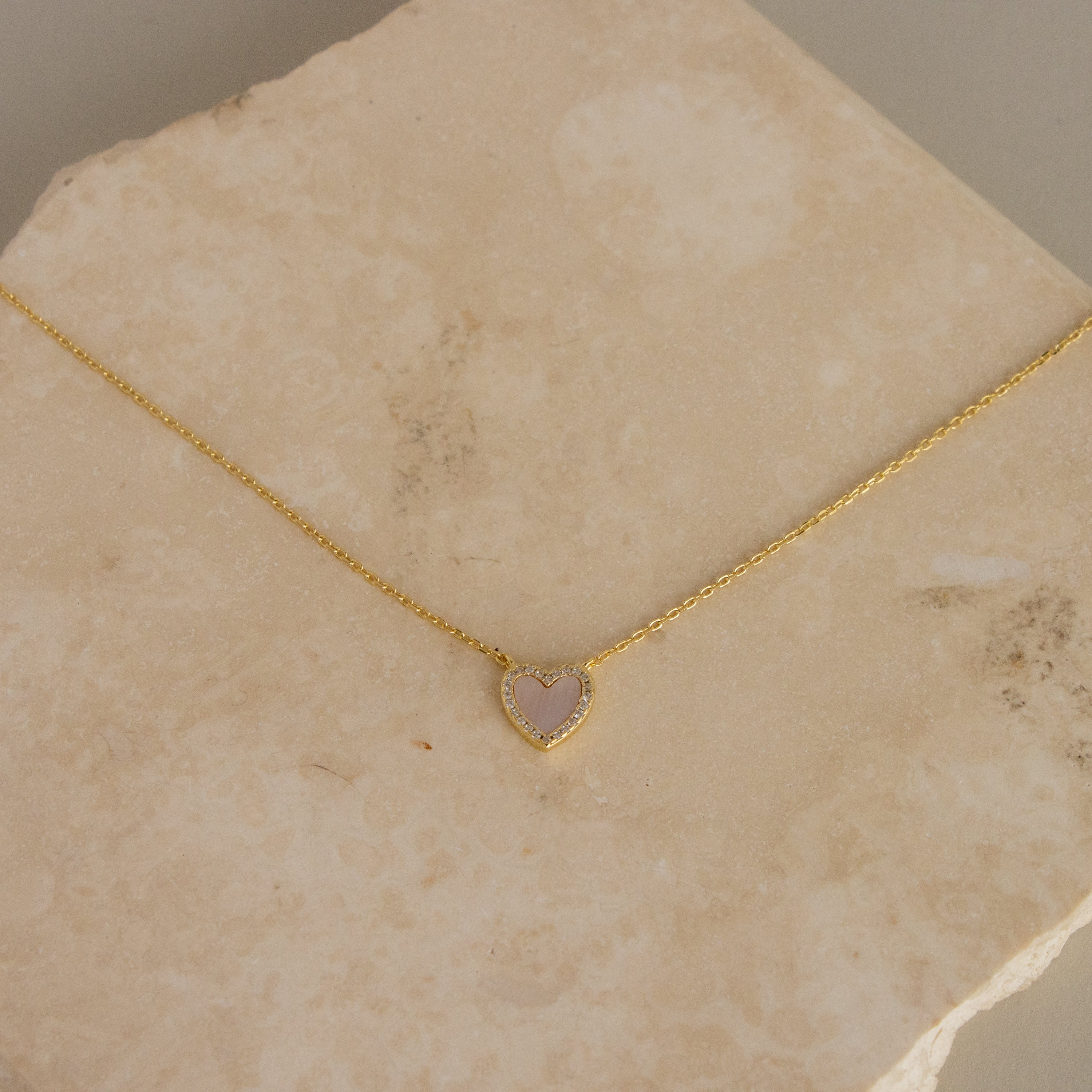 The Pink Pave Heart Necklace, featuring a sparkling heart-shaped pendant, is displayed on a beige stone surface.