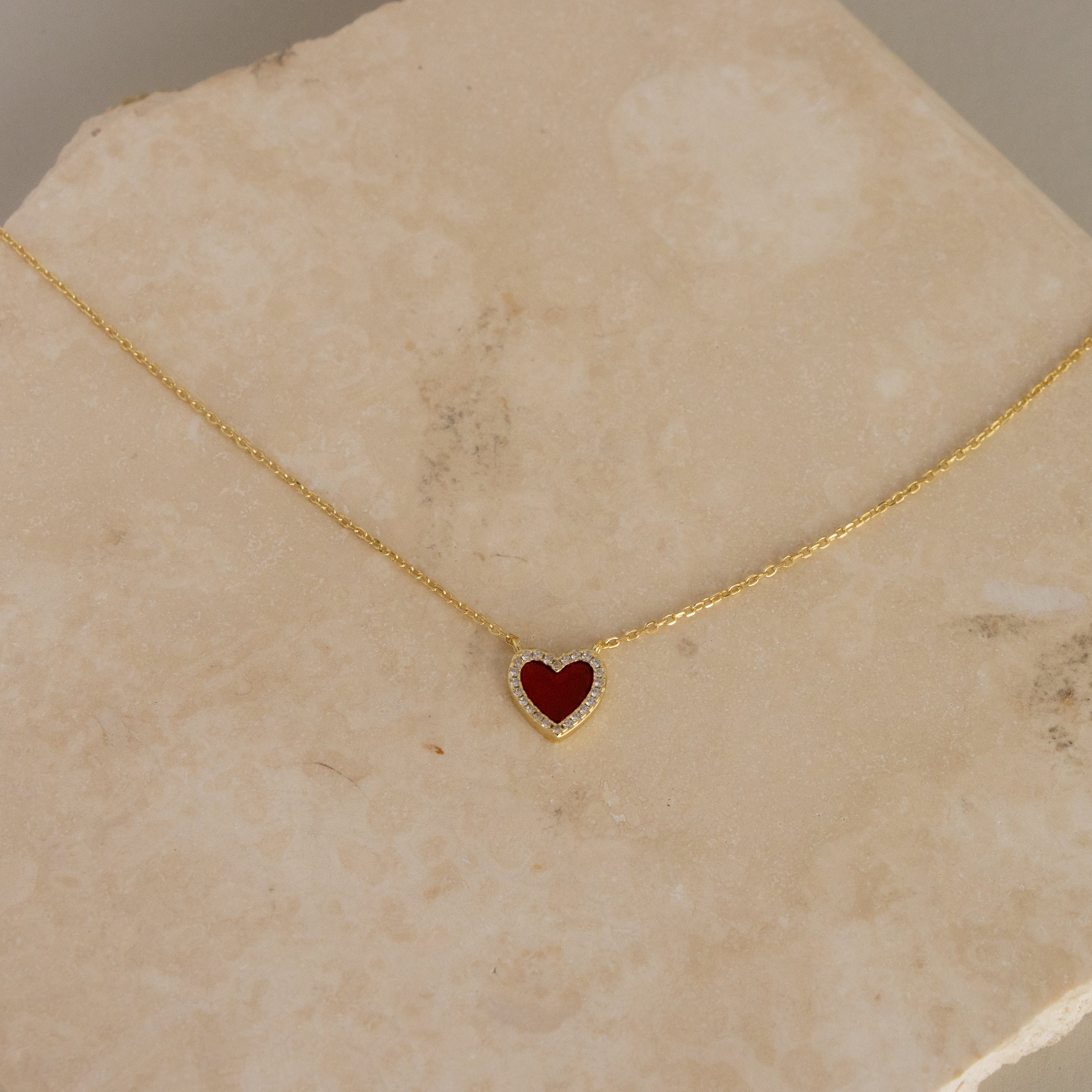Red Agate Pave Heart Necklace featuring a small red heart-shaped pendant, displayed on a beige stone surface.