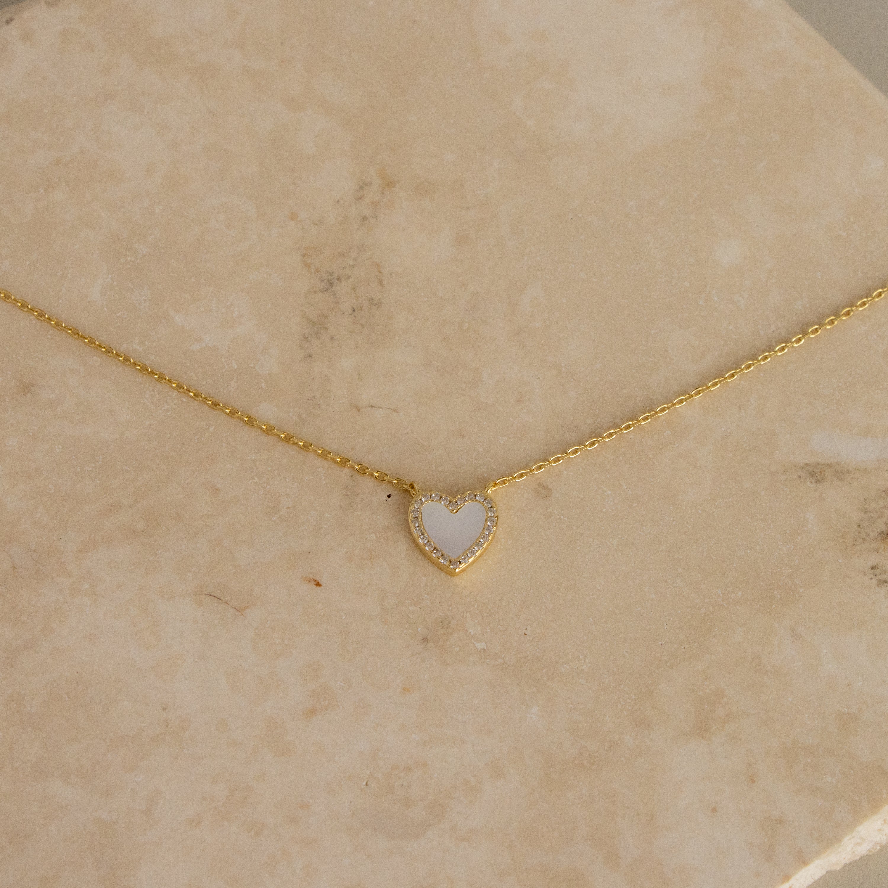 Mother of Pearl Pave Heart Necklace displayed on a beige marble surface, featuring a small white heart-shaped pendant.