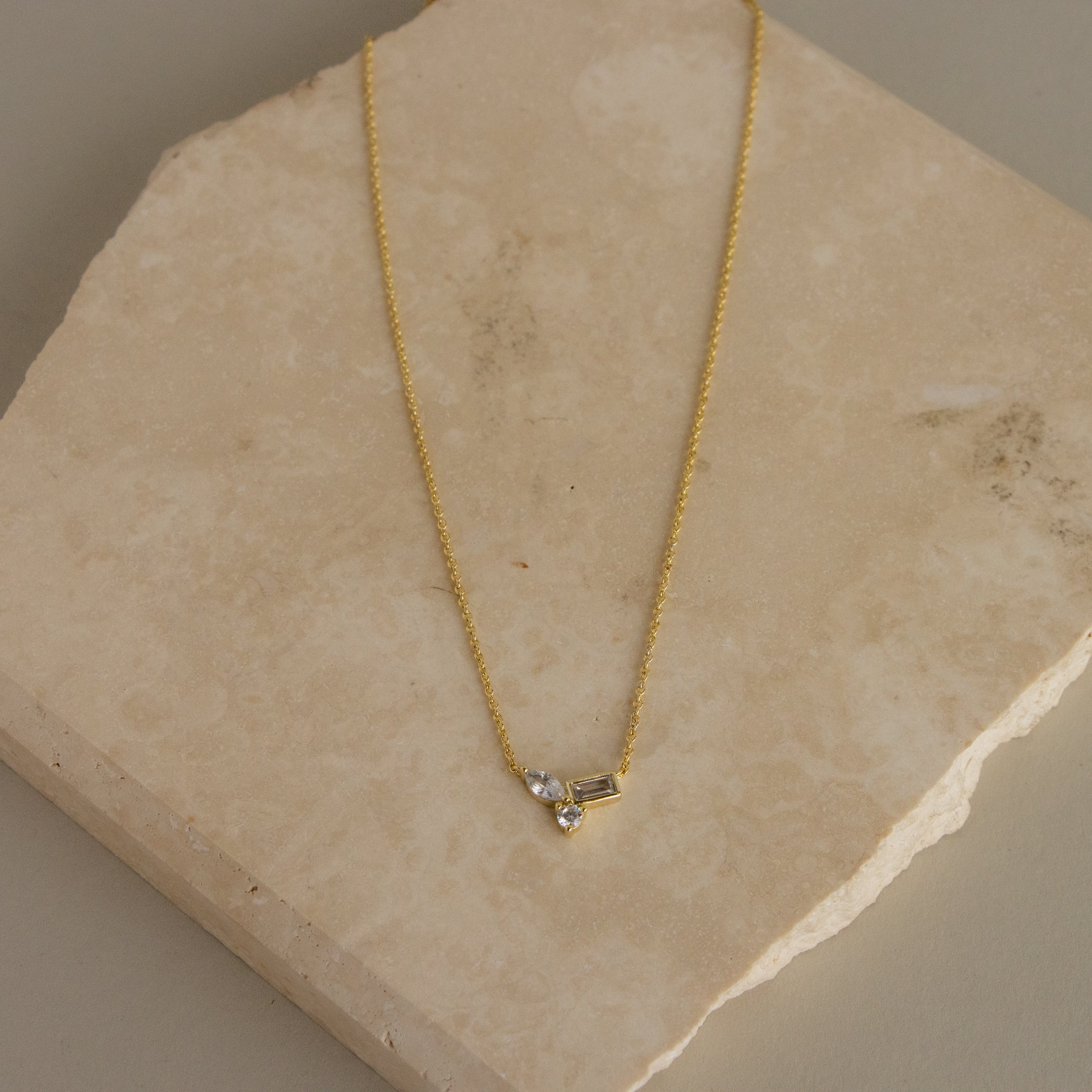 The Mixed Diamond Stone Charm Necklace features a small pendant with two clear stones, displayed on a beige stone surface.