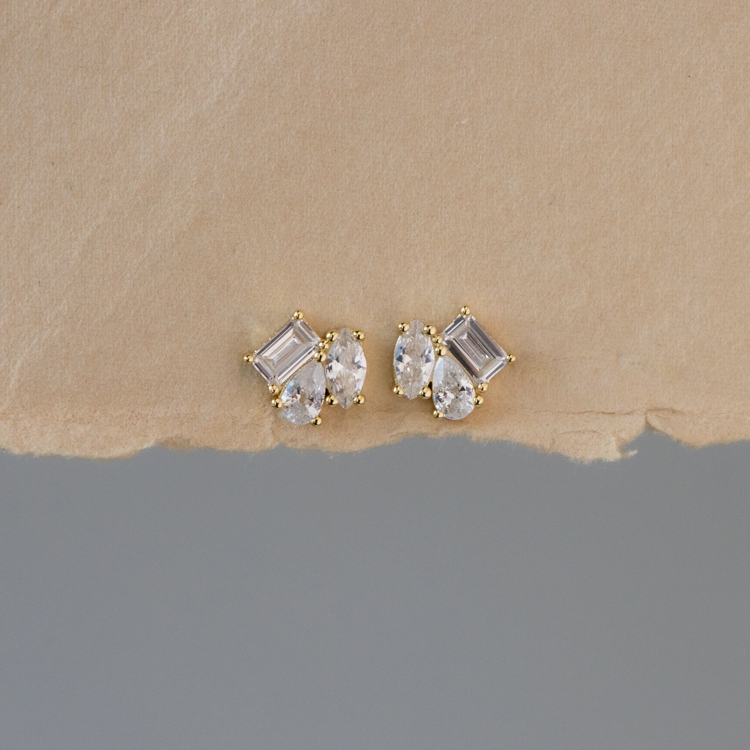 Mixed Diamond Cluster Studs featuring three clear gemstones each, shown on torn beige paper with a gray background.