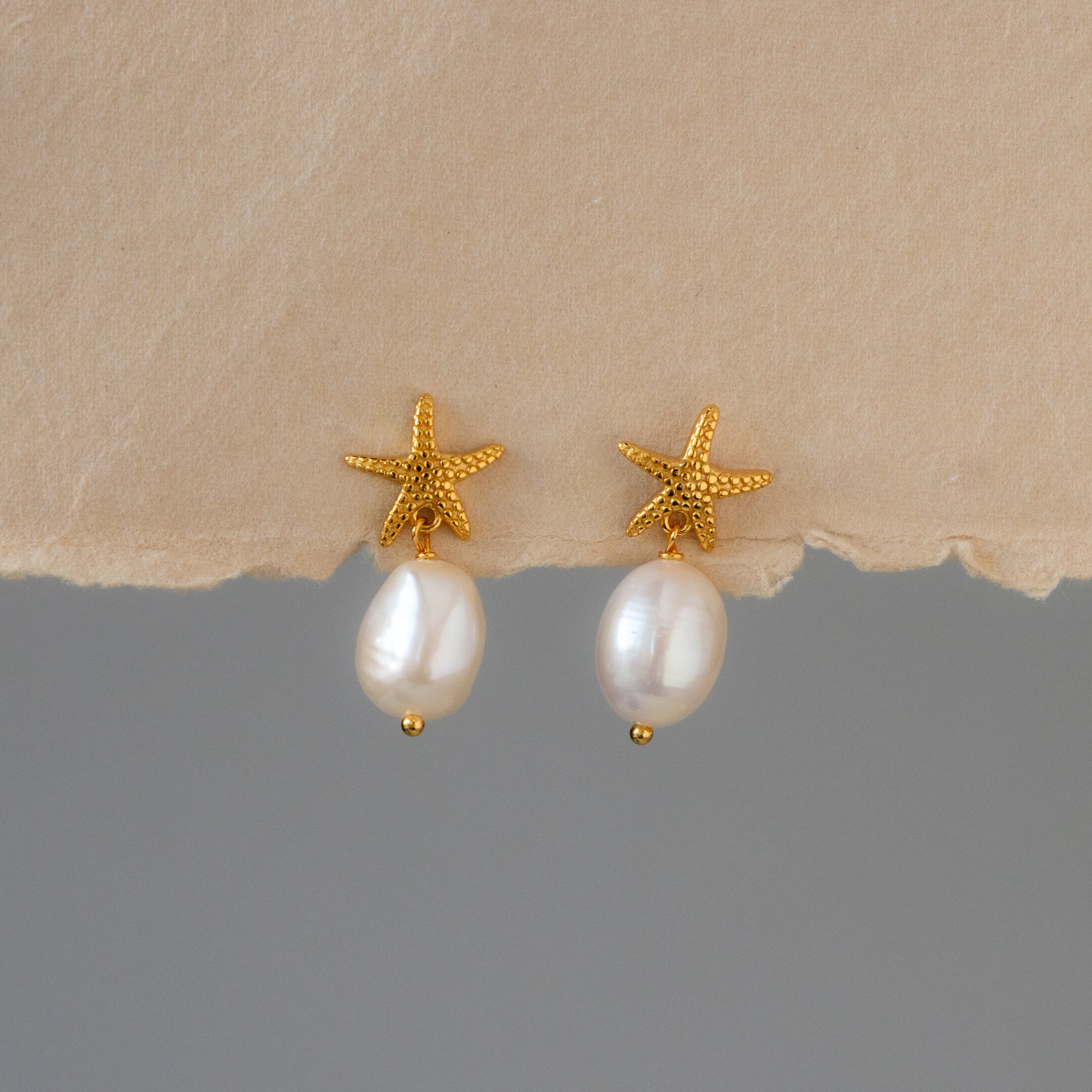 Starfish Baroque Pearl Drop Studs featuring gold starfish with dangling white pearls, displayed on a beige textured surface.