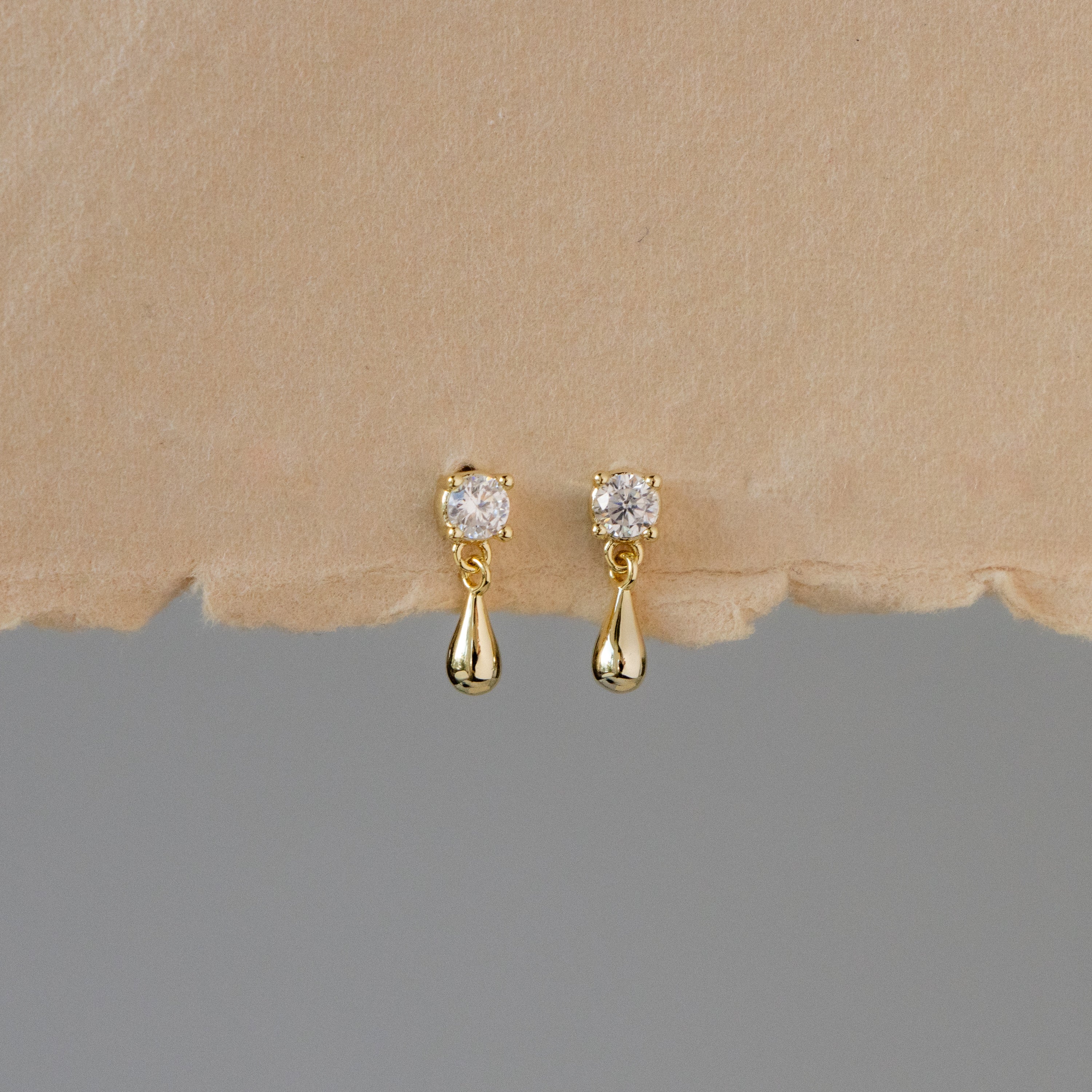 Mini Dangling Teardrop Diamond Studs with sparkling stones displayed on a beige textured background.