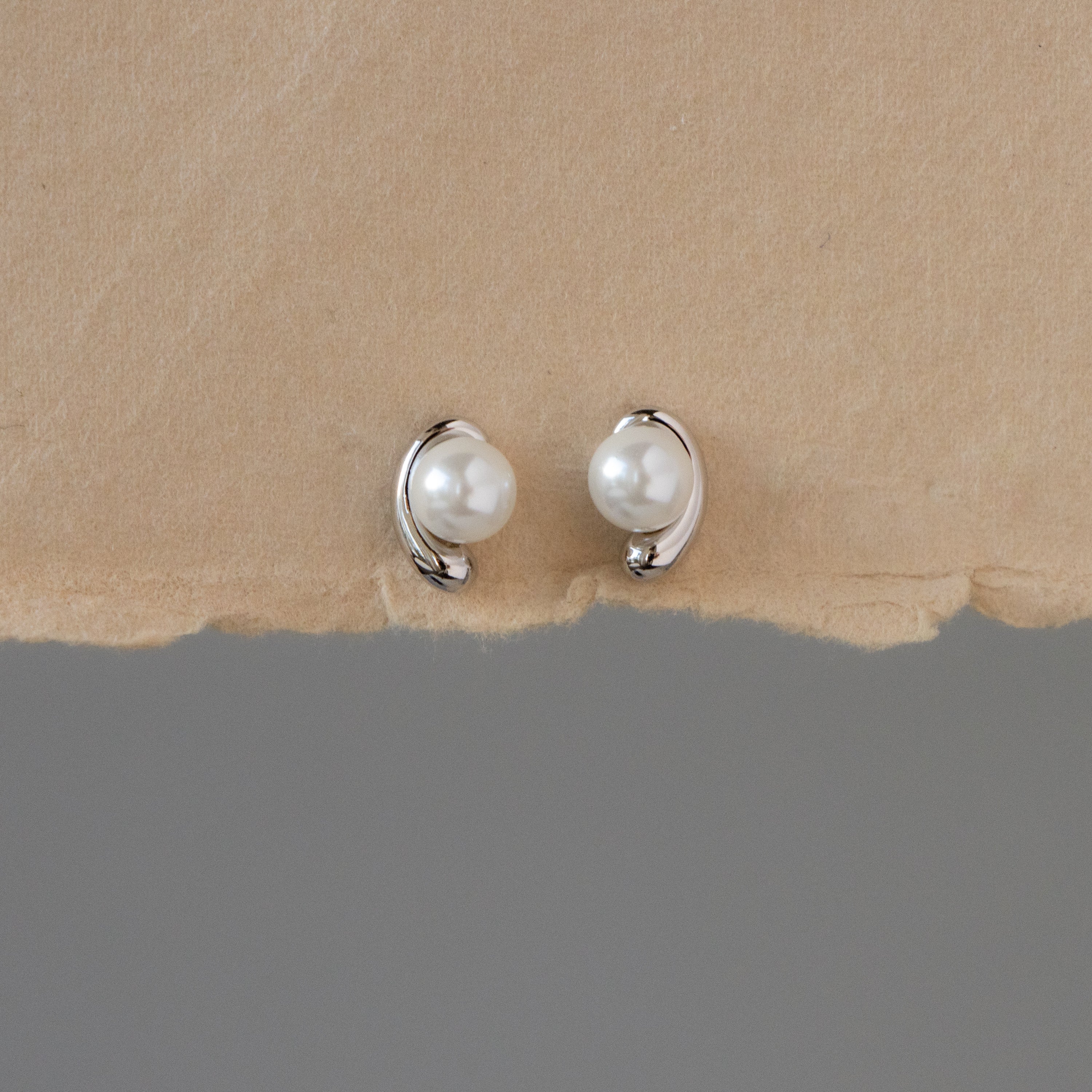 Silver Pearl Curve Studs with silver accents are displayed on a torn piece of beige paper.