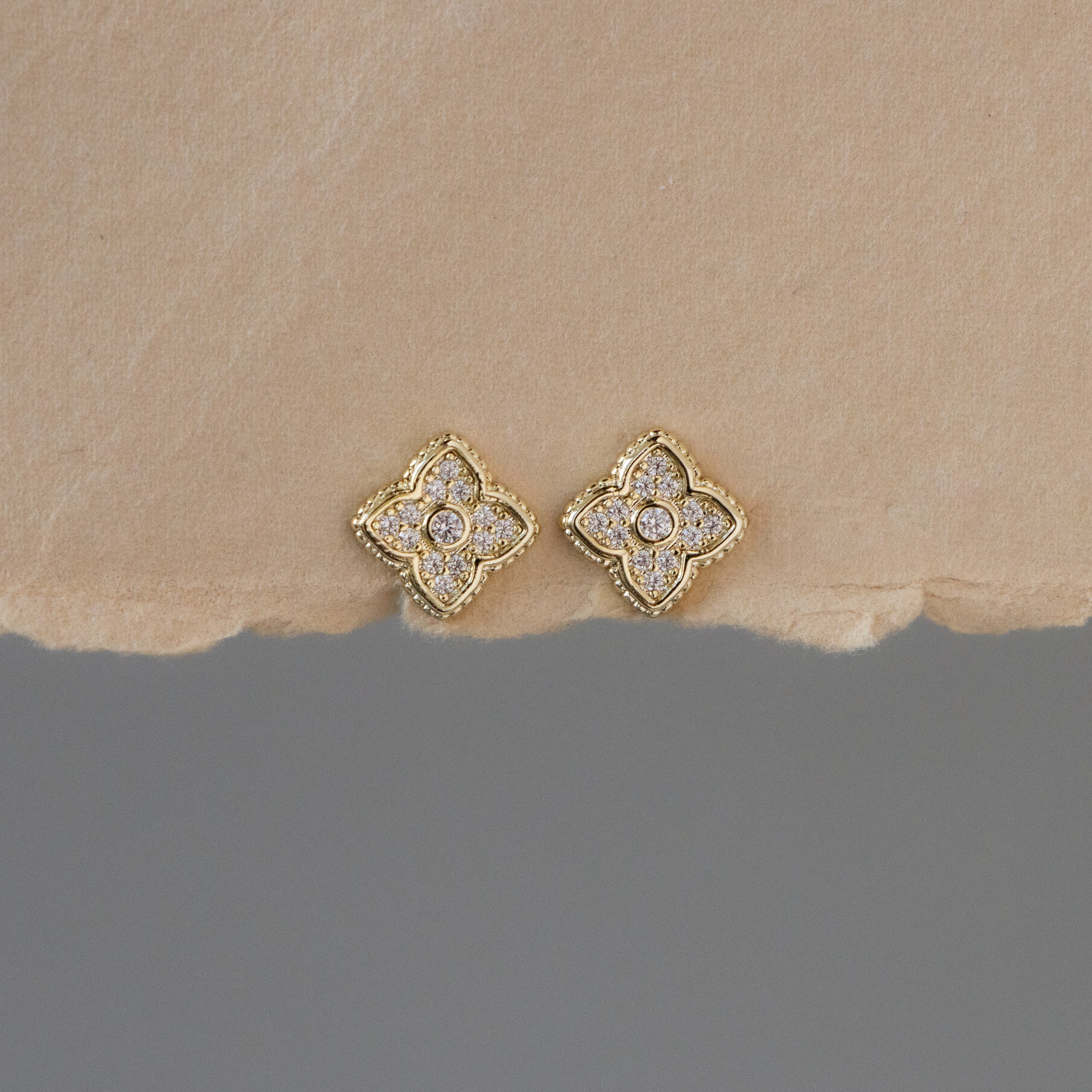 Bezel Pave Flower Studs—gold, clover-shaped stud earrings with small diamonds—displayed on a textured beige surface.