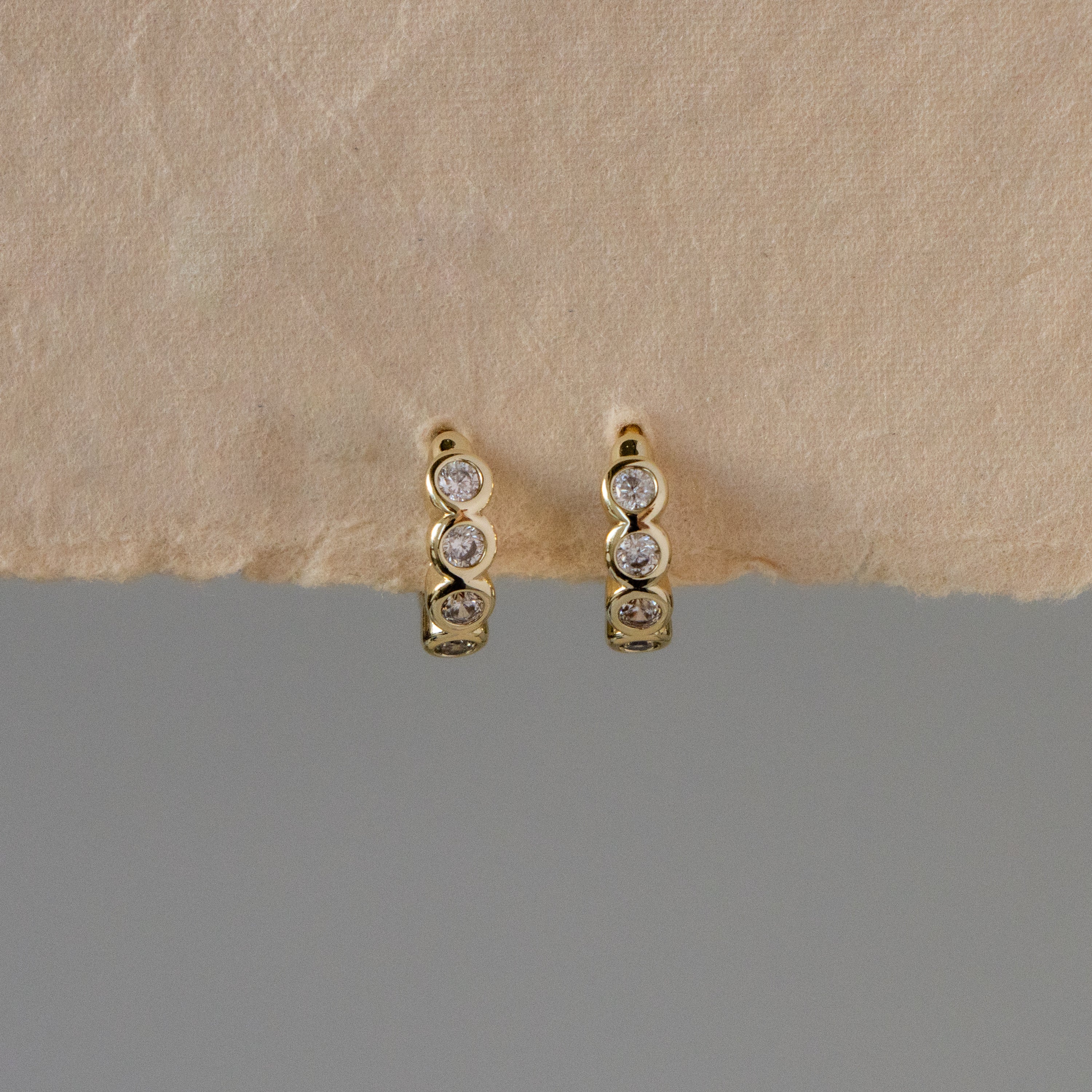 Mini Diamond Bezel Huggies featuring three round diamonds per earring, shown on a textured beige surface with a gray background.