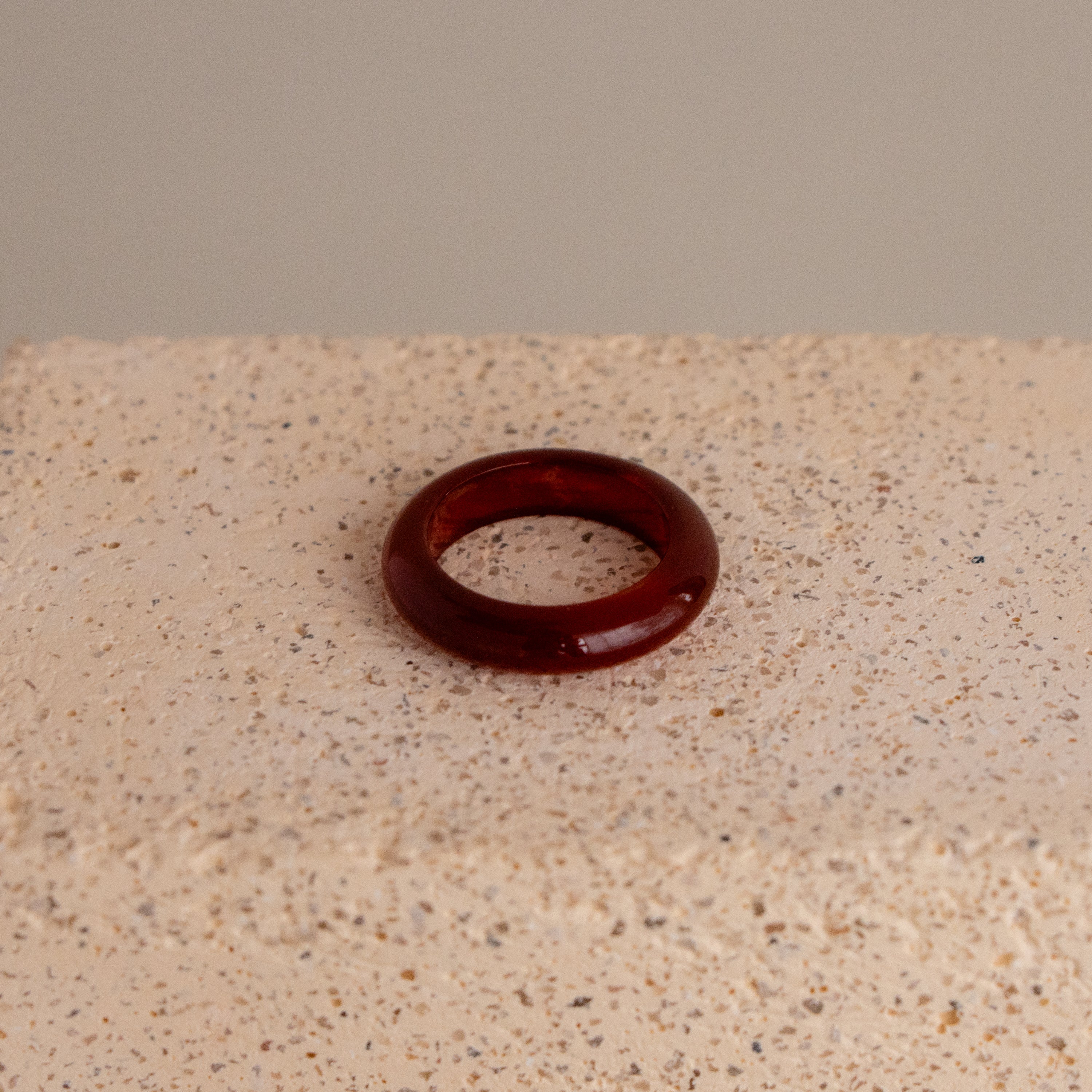 The Red Agate Halo Ring features a glossy, dark red finish and is displayed on a textured, beige stone surface.