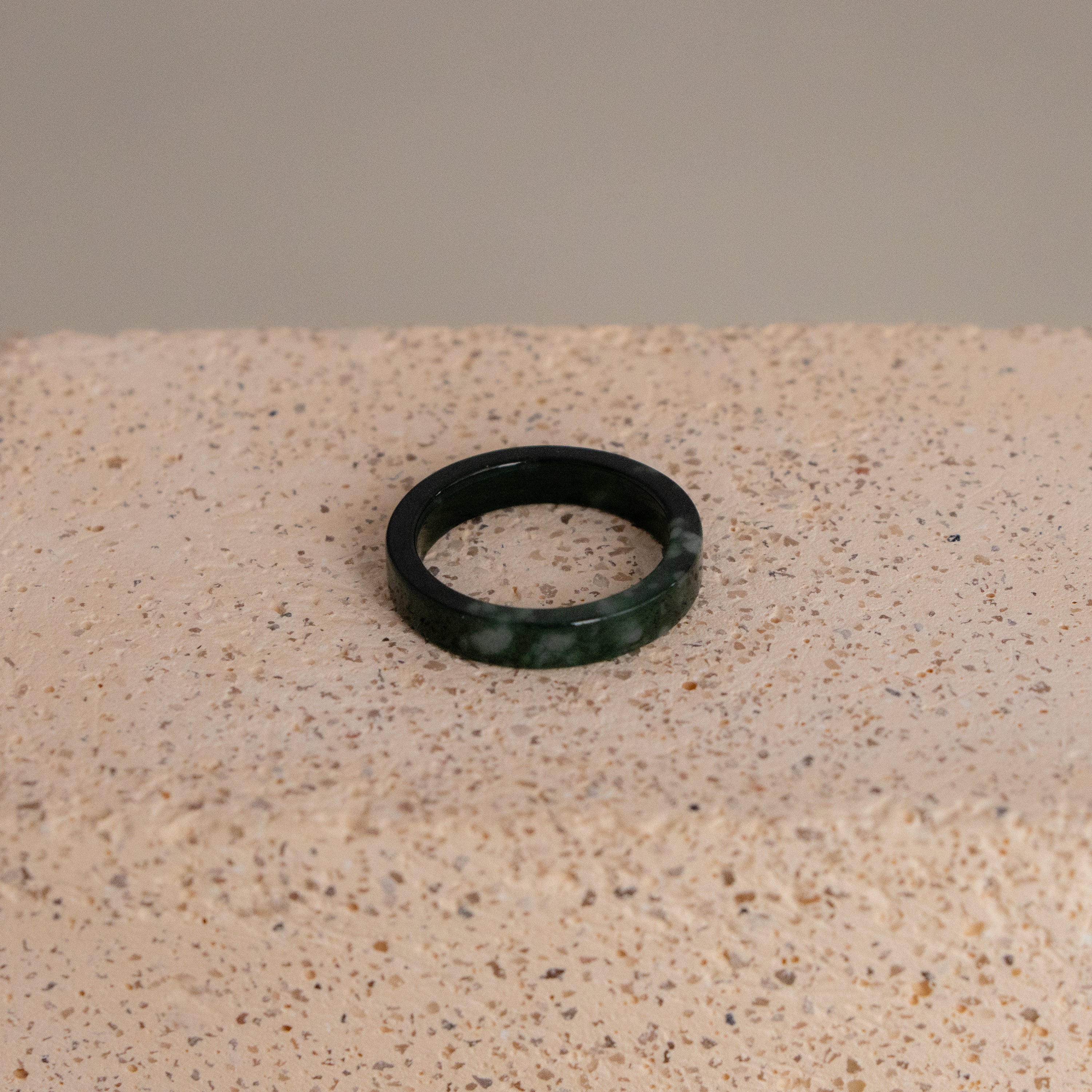 The Thick Dark Green Jade Ring is displayed on a textured, light beige stone surface.