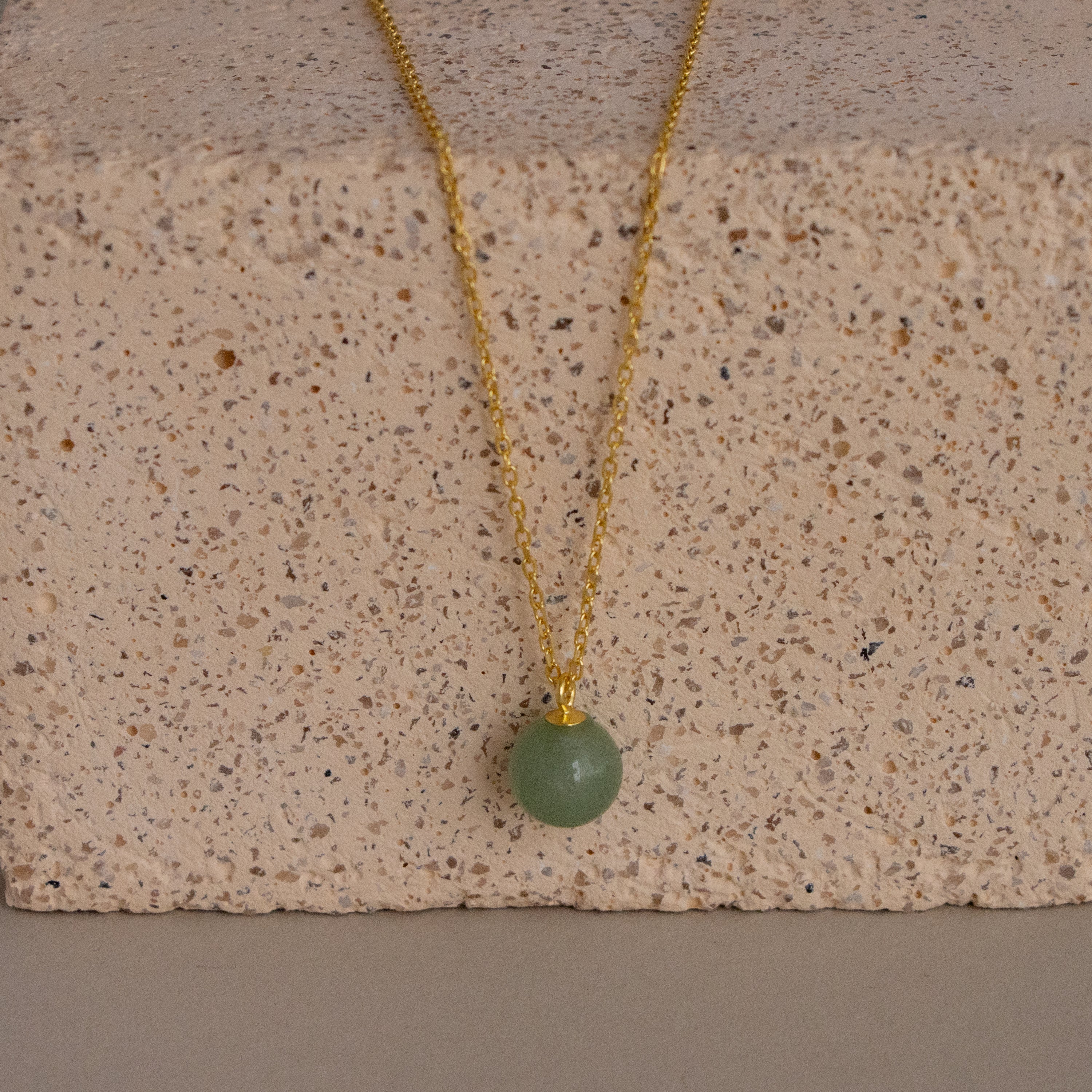 The Aventurine Bead Pendant Necklace features a gold chain with a round green stone pendant, displayed on a textured light-colored stone block.