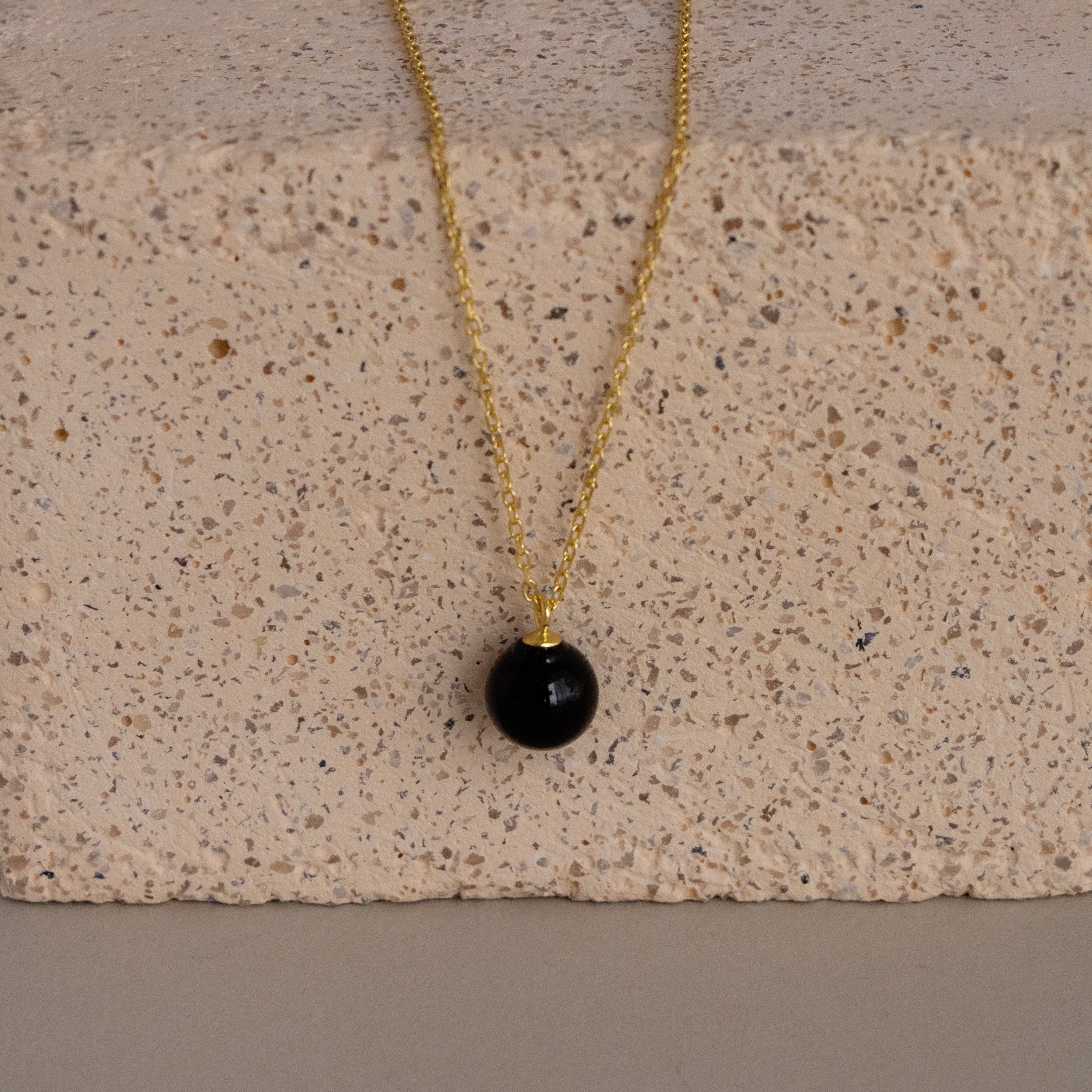 Onyx Bead Pendant Necklace featuring a round black onyx bead displayed against a textured, light-colored stone block.
