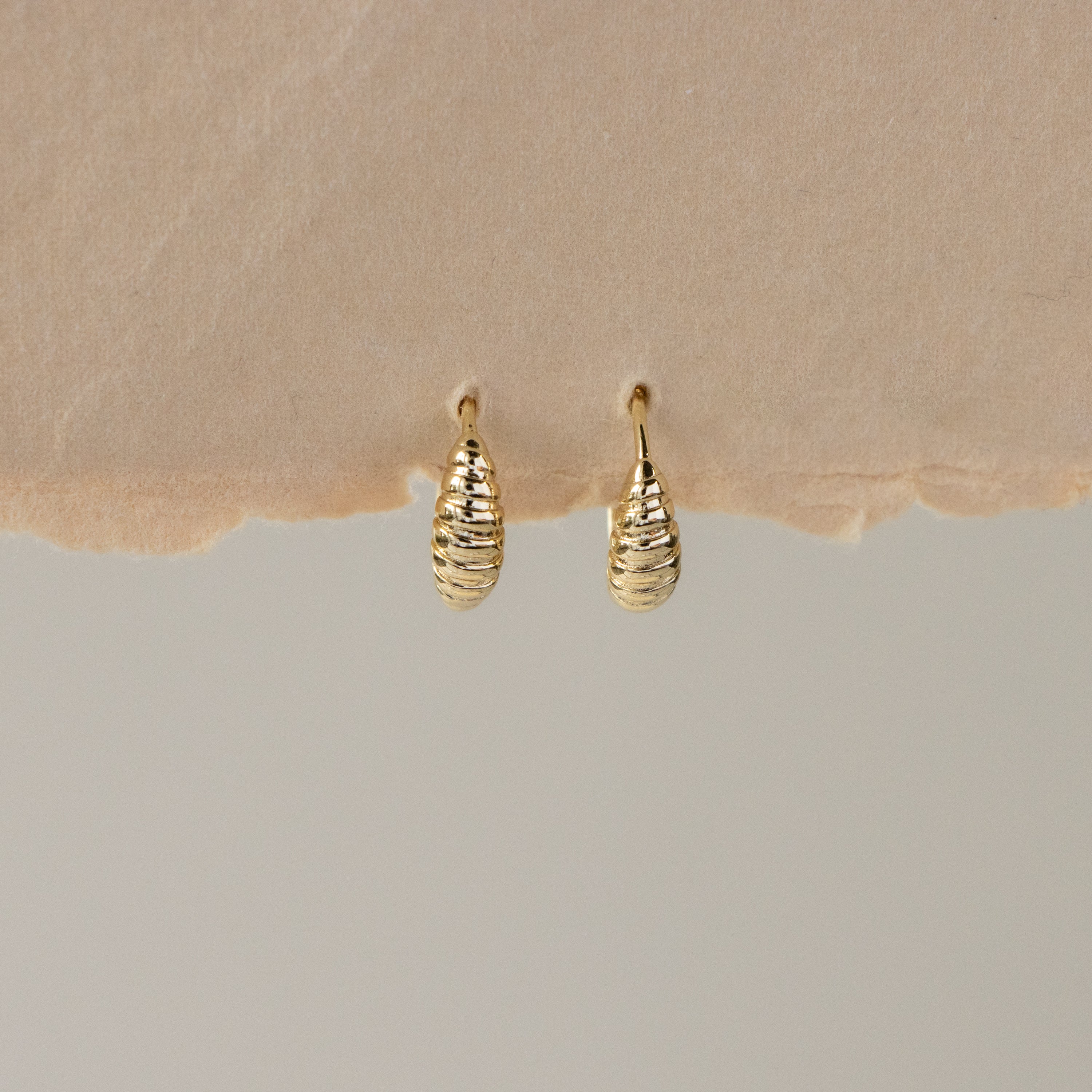 Dainty Croissant Huggies—small gold earrings with a textured, teardrop-inspired design—displayed on torn paper.