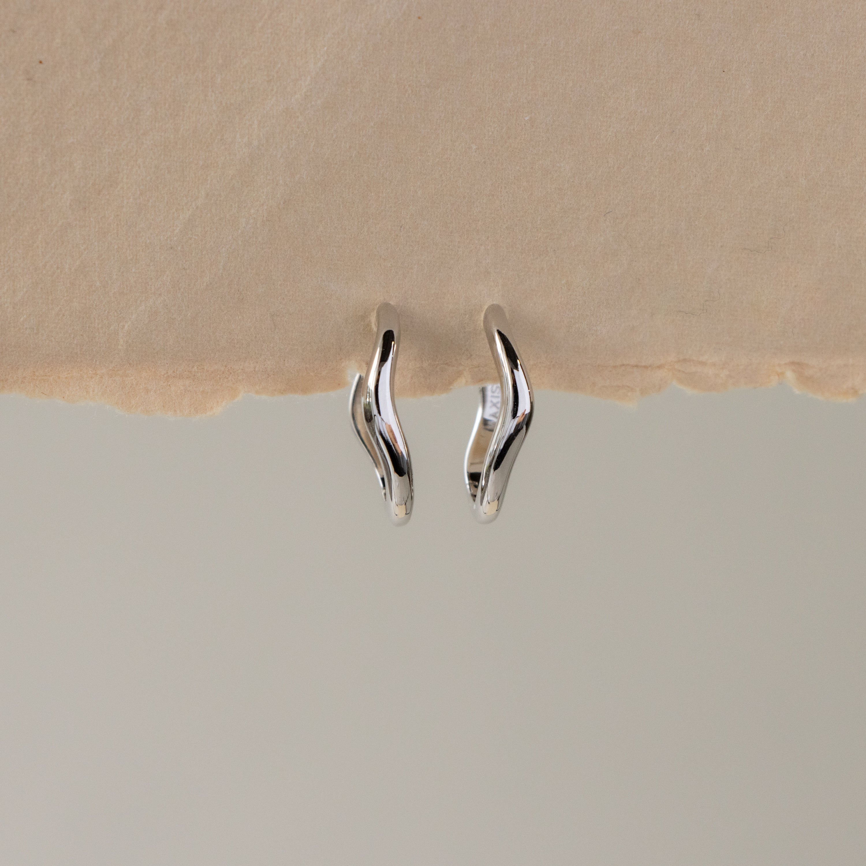 Dainty Silver Wave Huggies are shown on a textured beige surface.