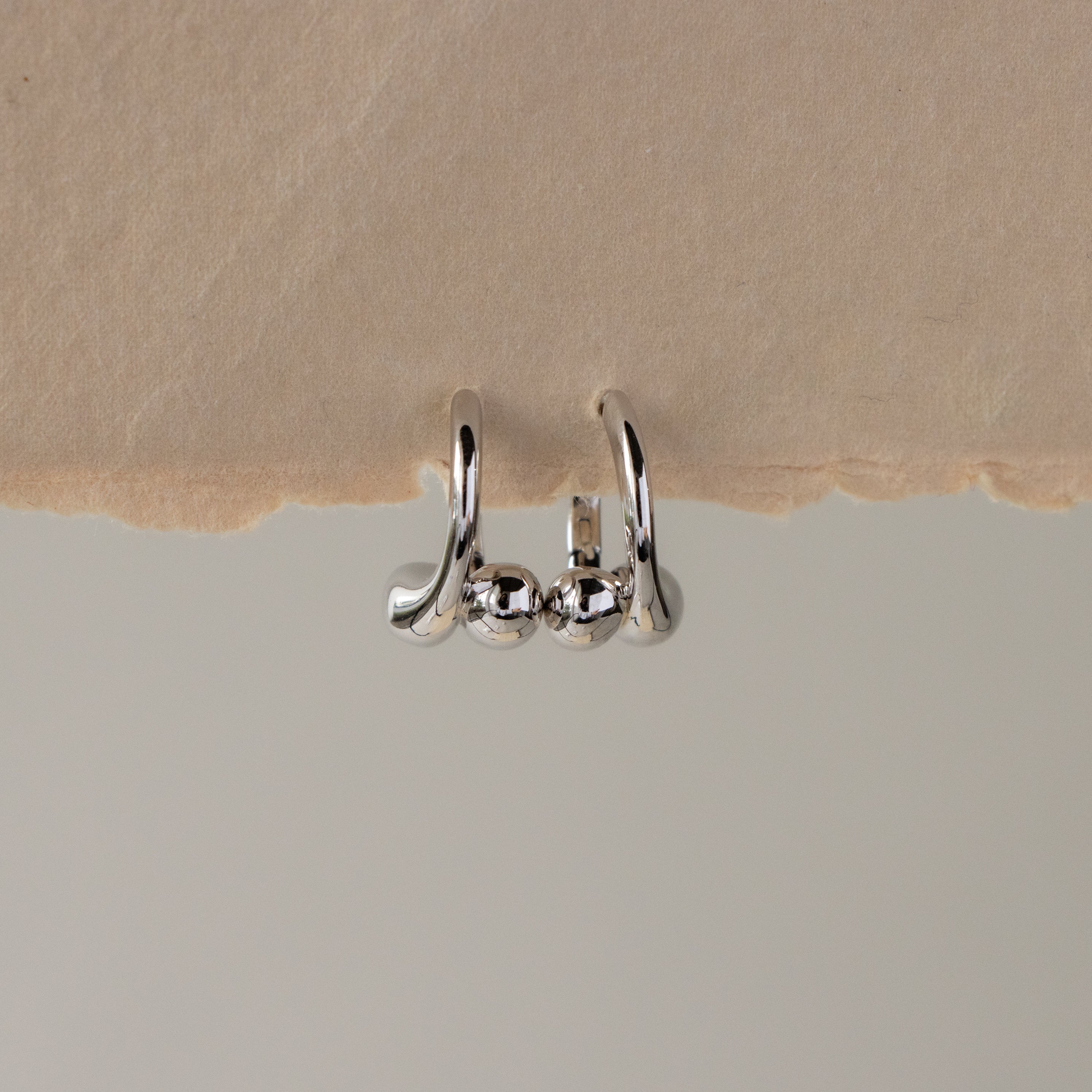 Silver Ball Curve Huggies, featuring three rounded beads, displayed on the edge of textured beige paper.