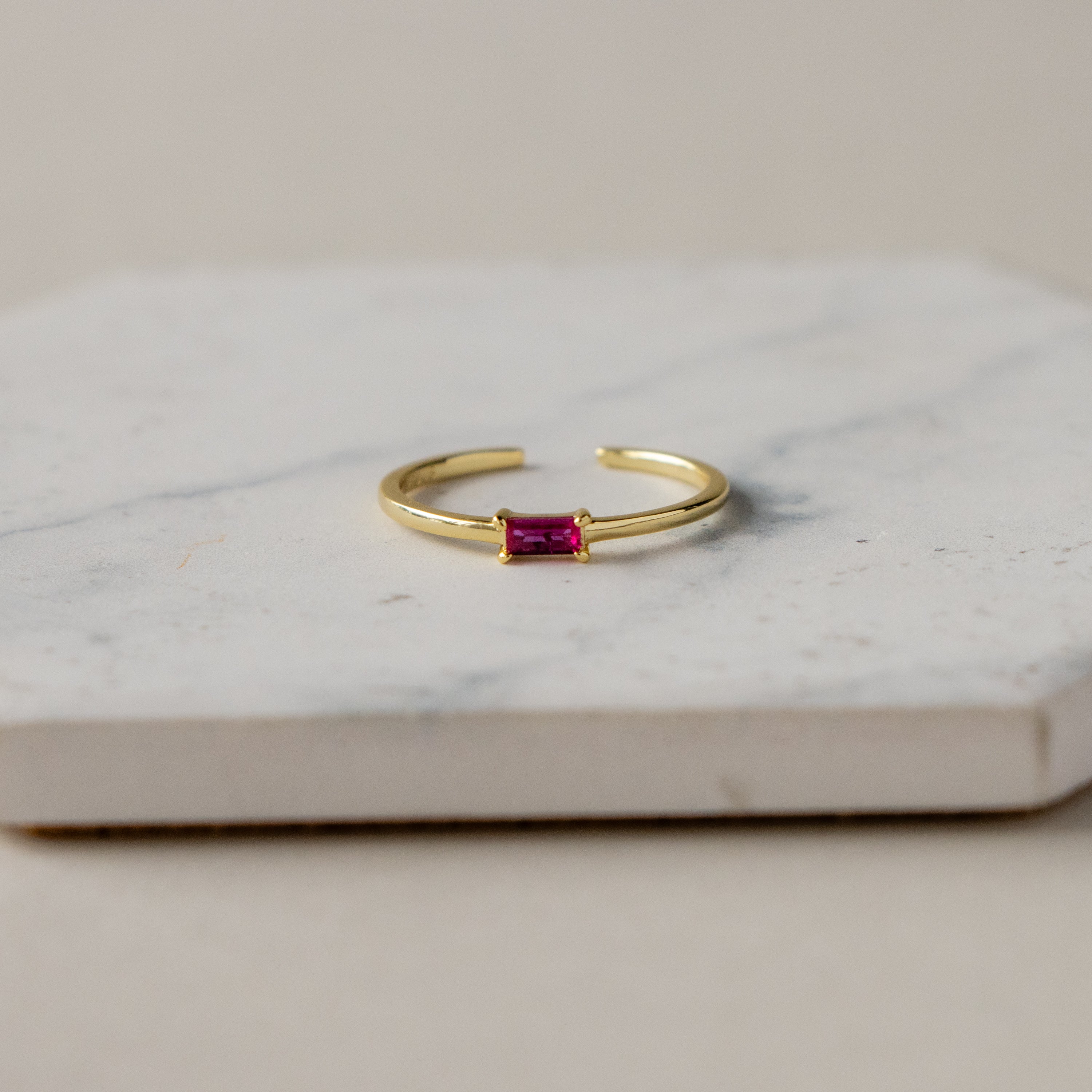 The Open Ruby Baguette Ring, featuring a rectangular pink gemstone, is displayed on a white marble surface.
