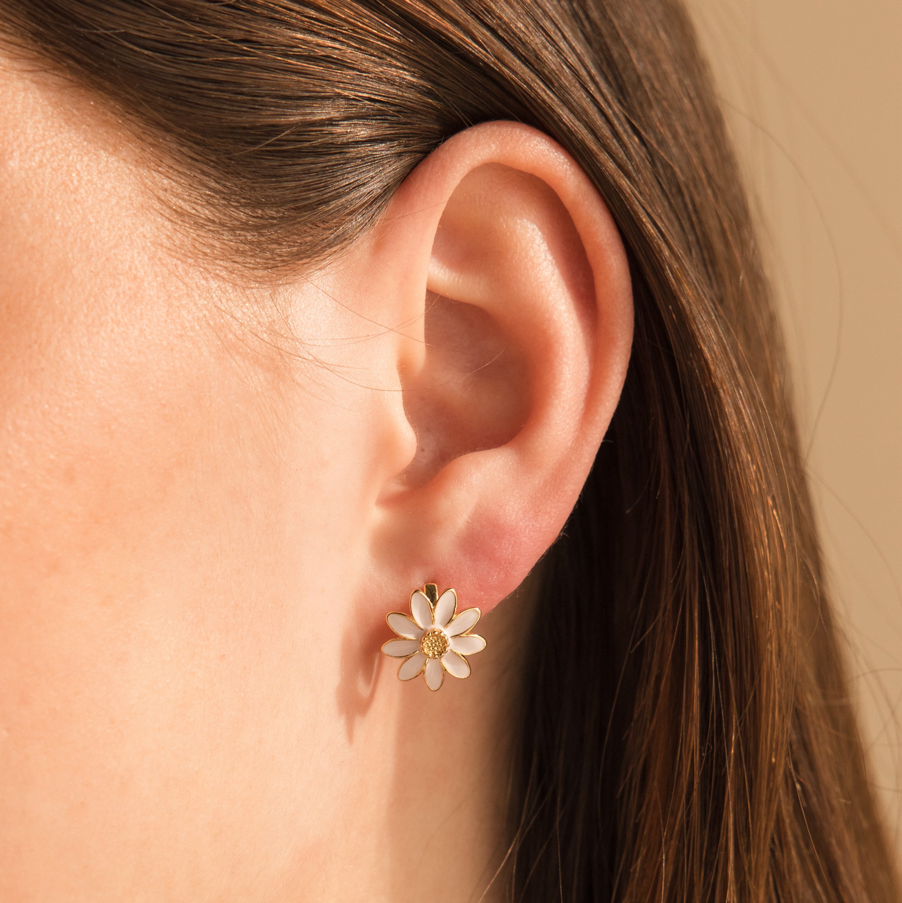 Close-up of a woman's ear wearing Daisy Huggies—white daisy-shaped studs ideal for minimalist jewelry lovers—with brown hair partially visible.