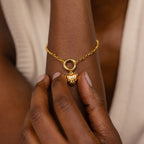 A hand touches a gold bracelet featuring the Vintage Heart Charm in 18K Gold on a wrist with a cream top—an elegant addition to any jewelry collection.