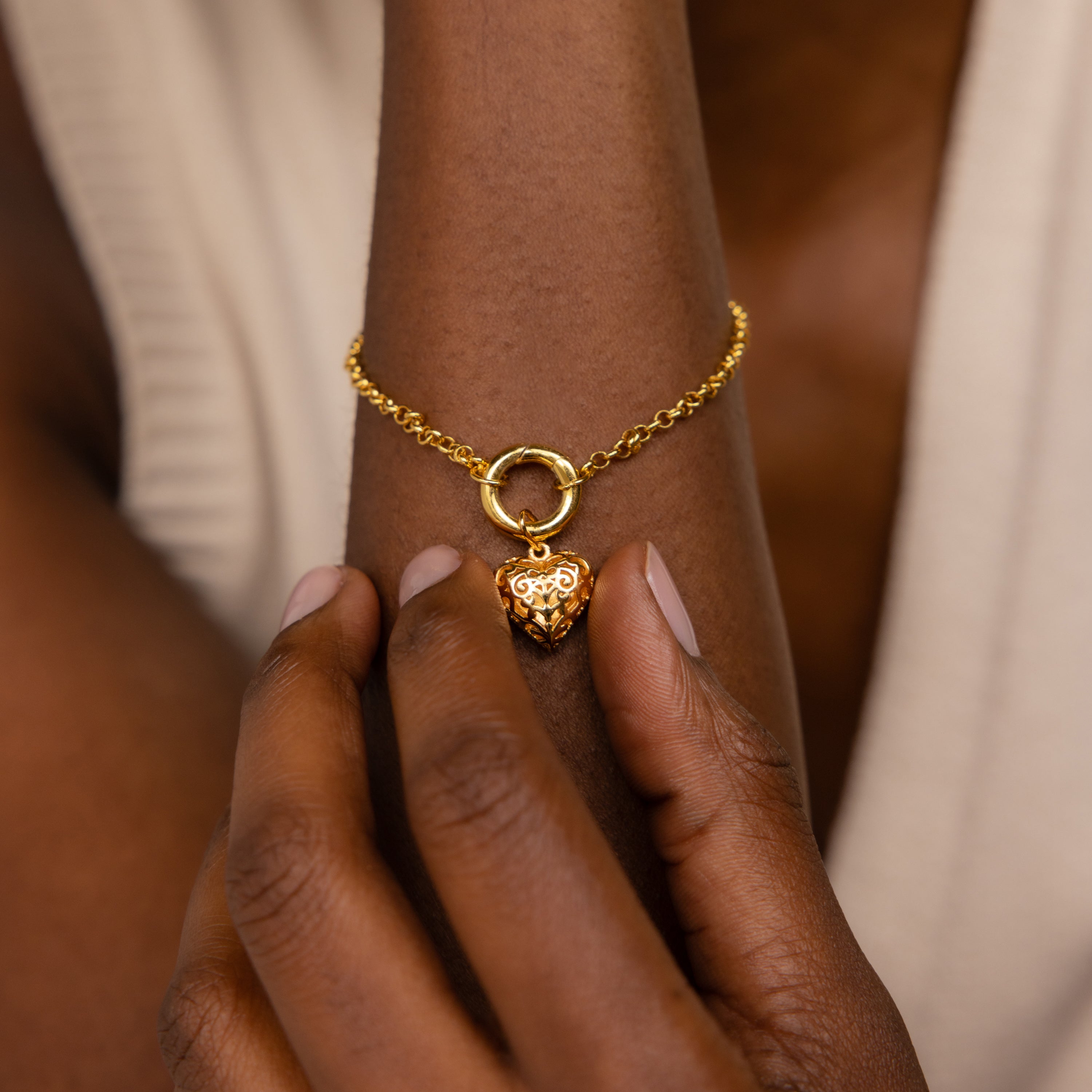 A hand touches a gold bracelet featuring the Vintage Heart Charm in 18K Gold on a wrist with a cream top—an elegant addition to any jewelry collection.