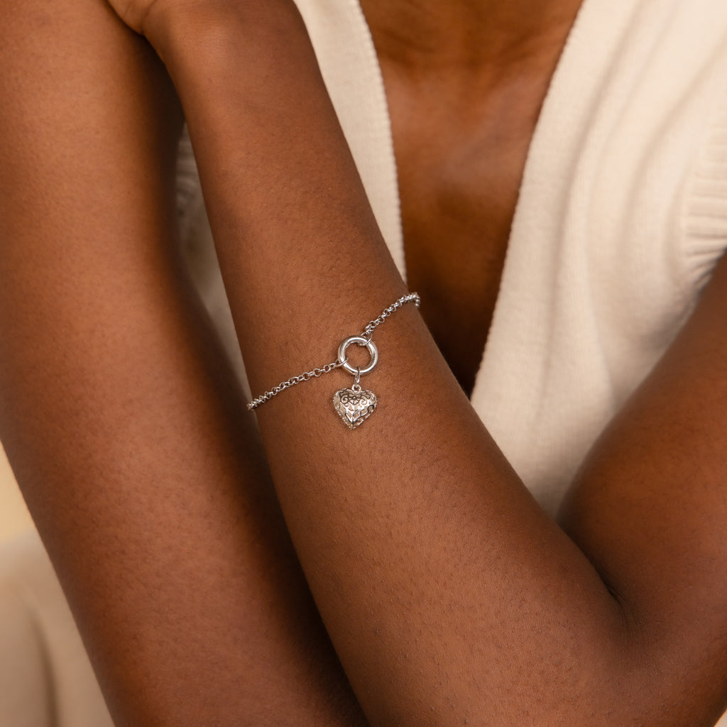 The Rolo Circle Charm Bracelet in White Gold, featuring a heart-shaped charm and detachable hinged circle, is shown on a wrist with a white sleeveless top.