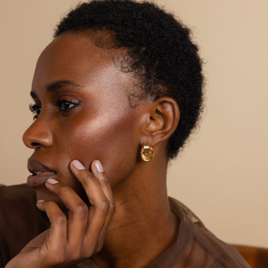 A woman with short curly hair wears Triple Knot Earrings in 18K Gold, touches her face, and gazes to the side.