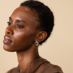 A woman with short curly hair, wearing Triple Knot Earrings and a sheer brown top, gazes left—making these earrings an inspiring anniversary gift.