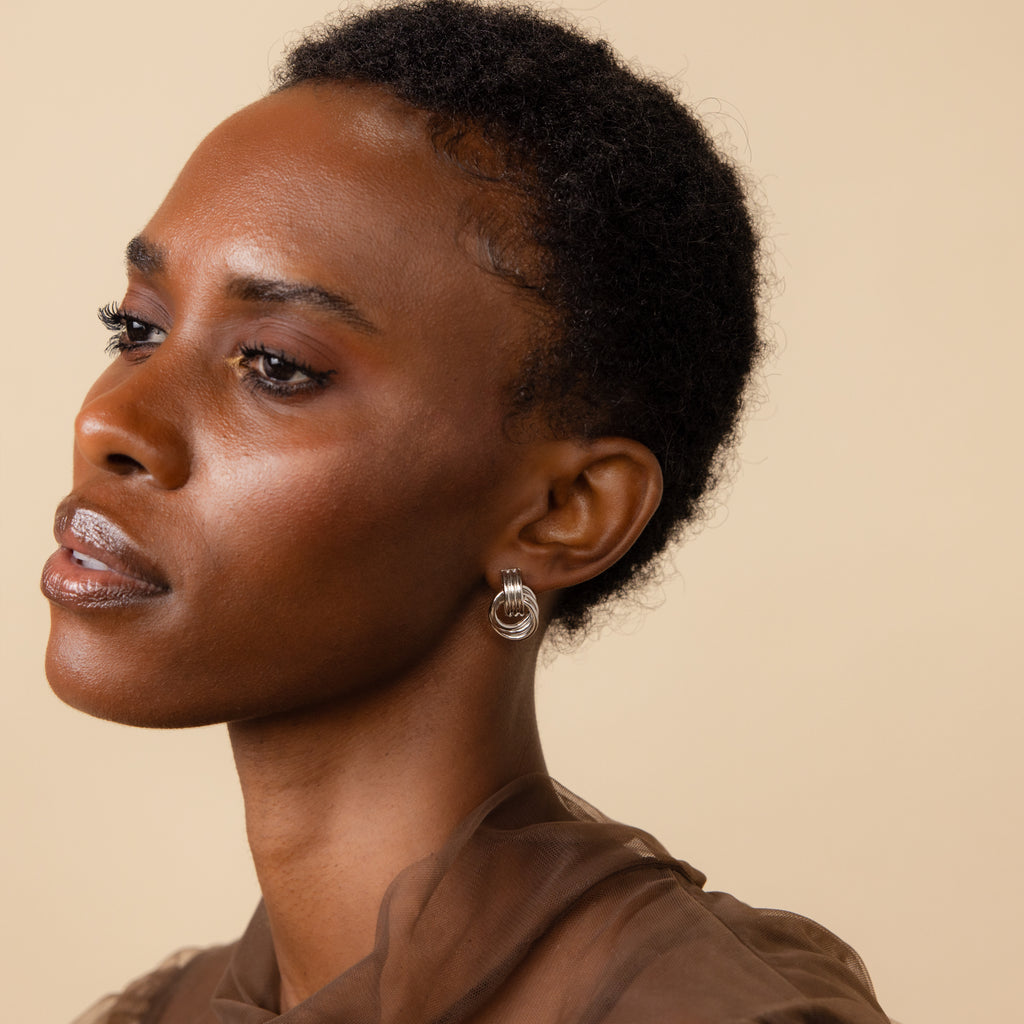 A woman with short curly hair wears the elegant Triple Knot Earrings in Silver and a sheer brown top—an ideal anniversary gift.