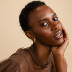A woman poses wearing the gold-toned Mini Triple Knot Earrings, resting her hand gently on her face