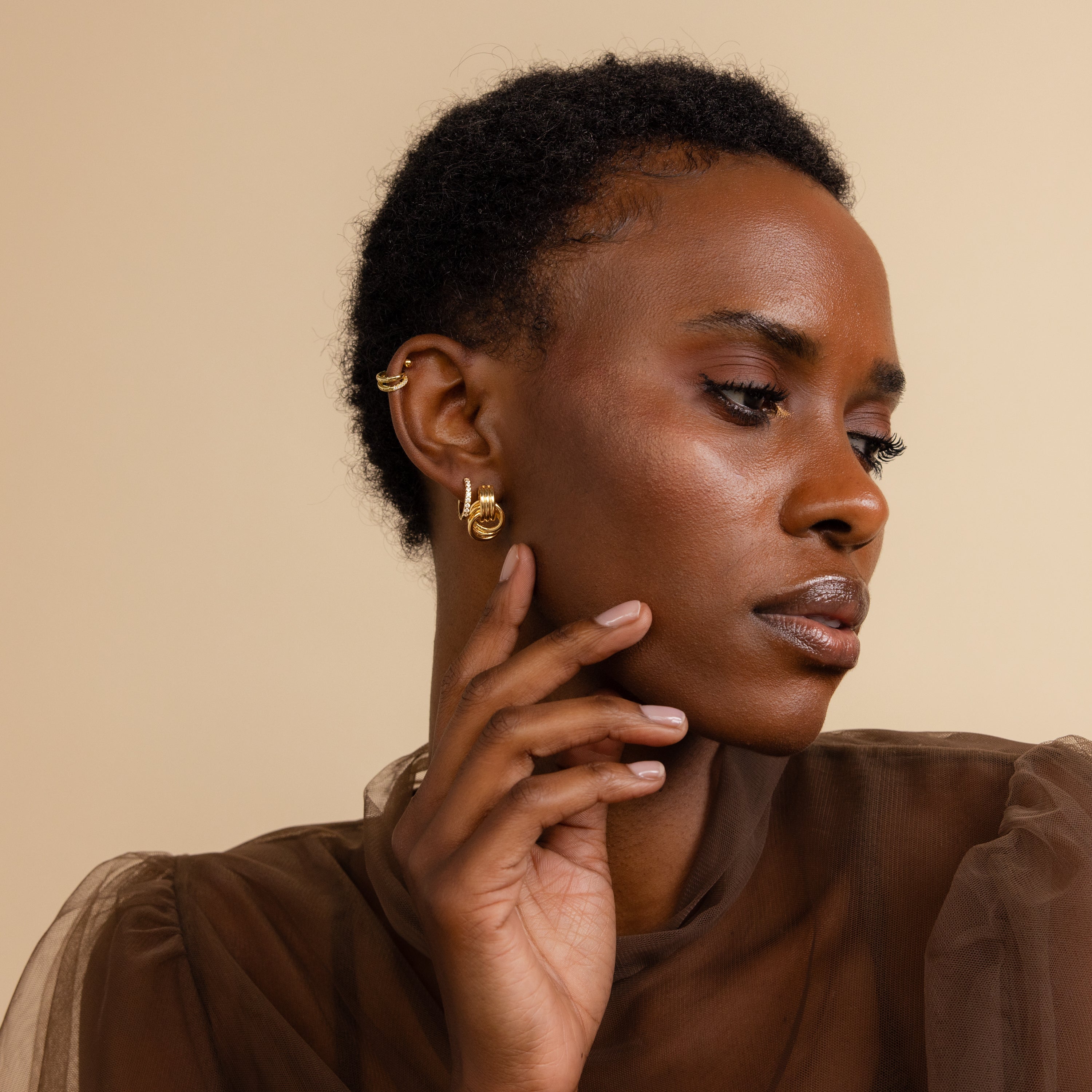 A model wears gold Mini Triple Knot Earrings along with other gold ear accessories, lightly touching her face in a side-profile pose