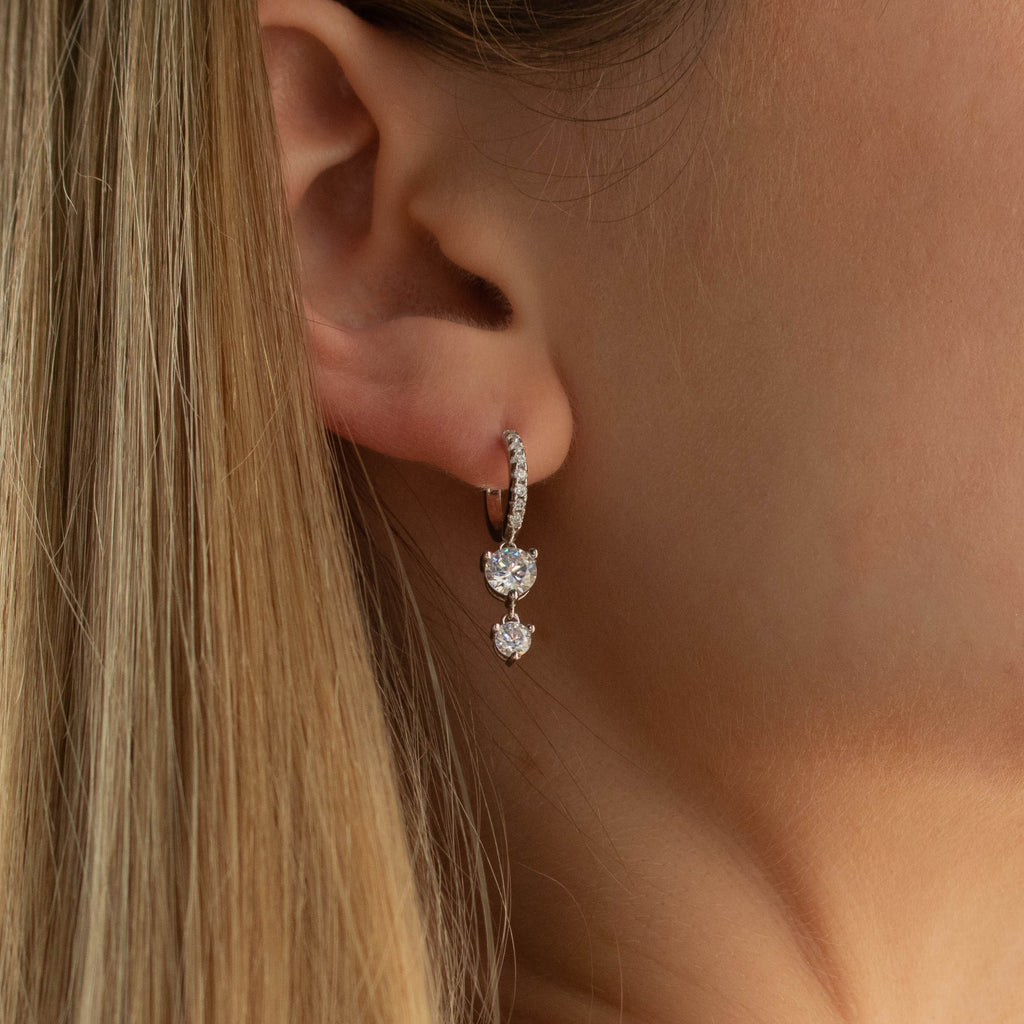 Close-up of a womans ear wearing the Caroline Pave Drop Huggies in Silver, featuring embellished hoops with two dangling round gemstones for a chic, modern style.