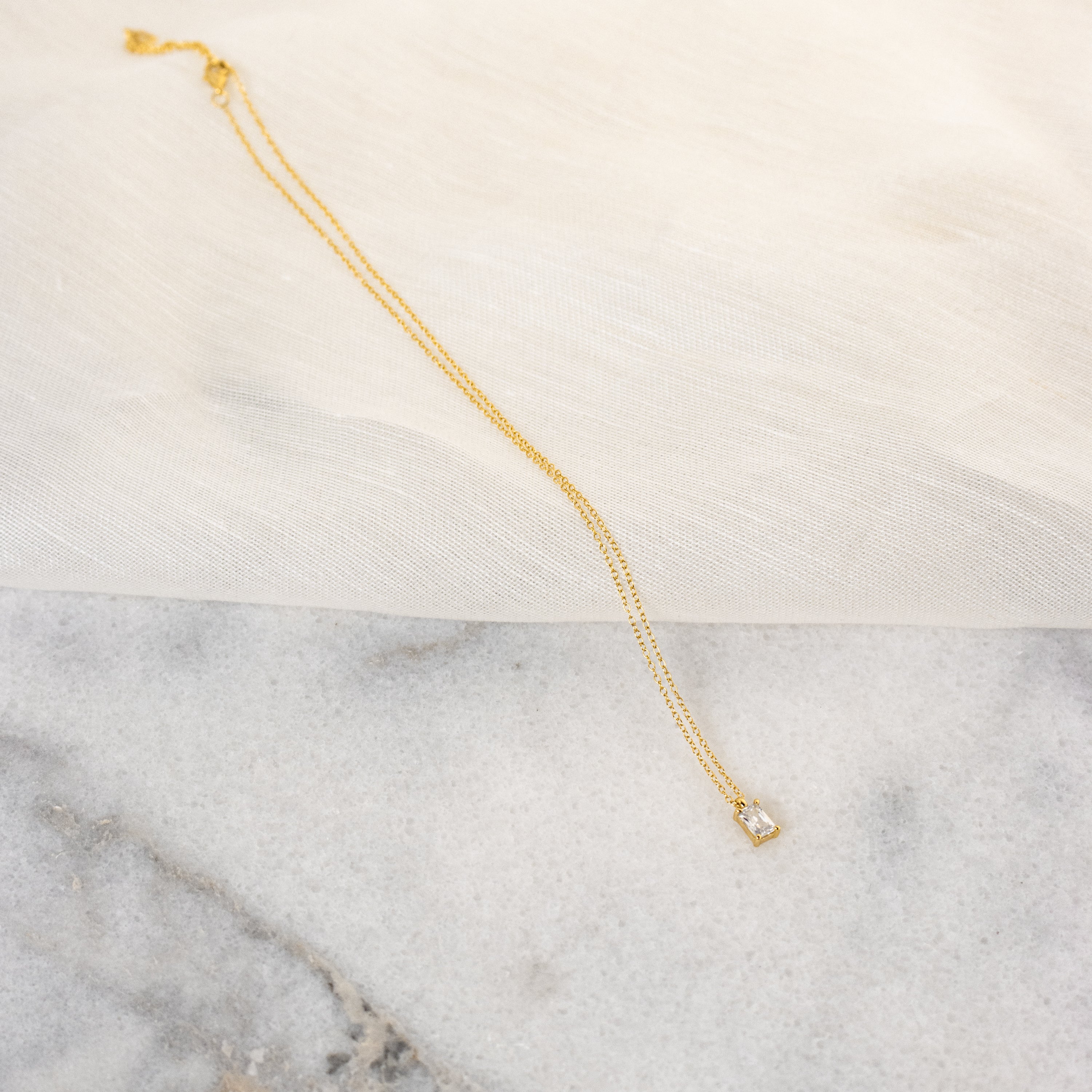 The Sage Diamond Necklace in 18K Gold features a delicate square pendant, radiating timeless elegance against a marble and fabric backdrop.