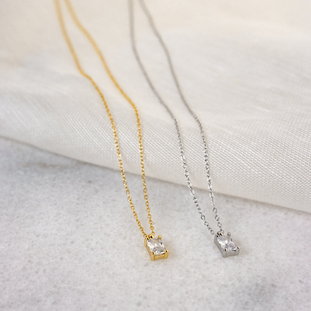 Two Sage Diamond Necklaces—one gold, one silver, each featuring a small rectangular pendant—rest on light fabric, showcasing the refined elegance of this luxury necklace collection.