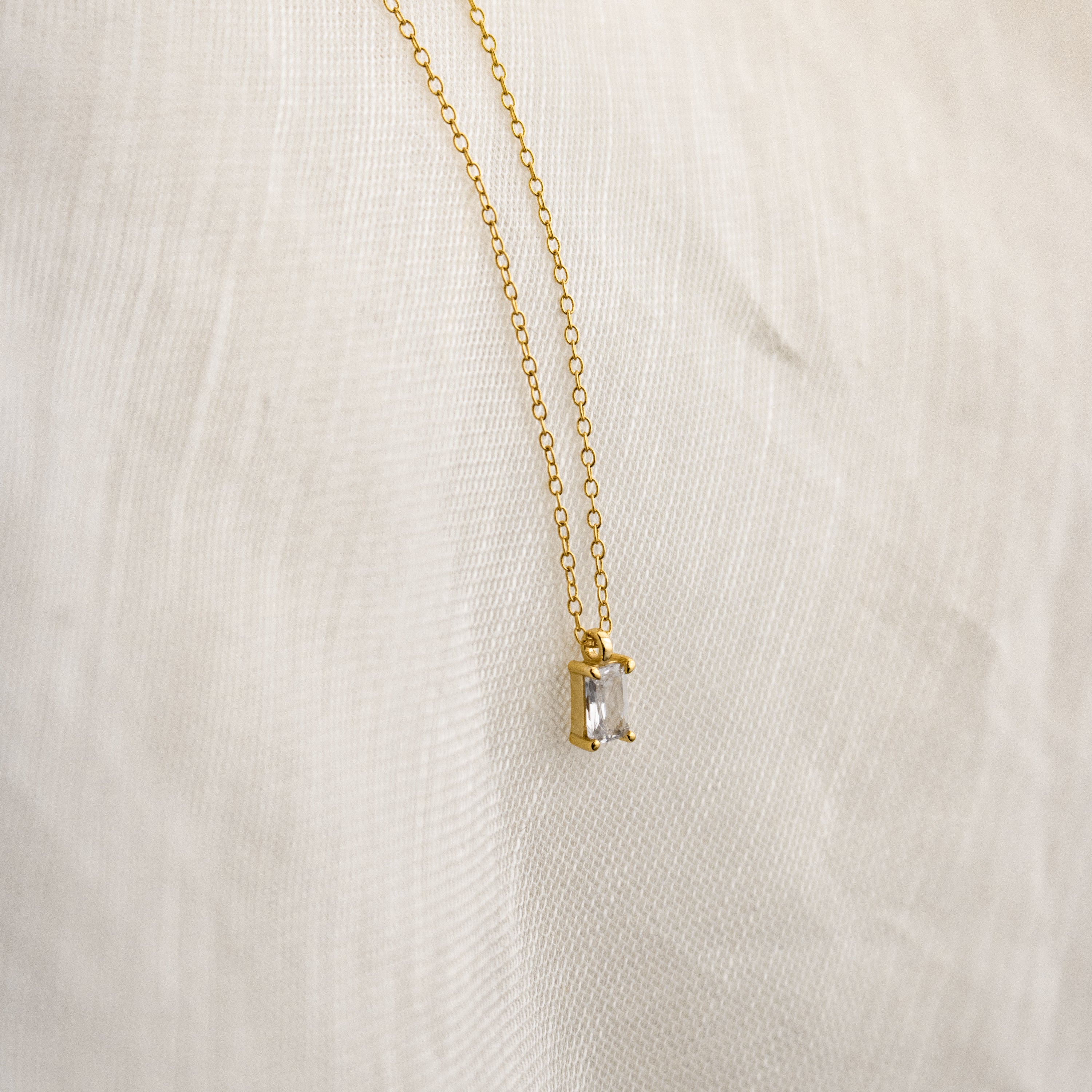 The Sage Diamond Necklace features a delicate gold chain with a rectangular clear gemstone pendant, elegantly displayed against a white fabric background.