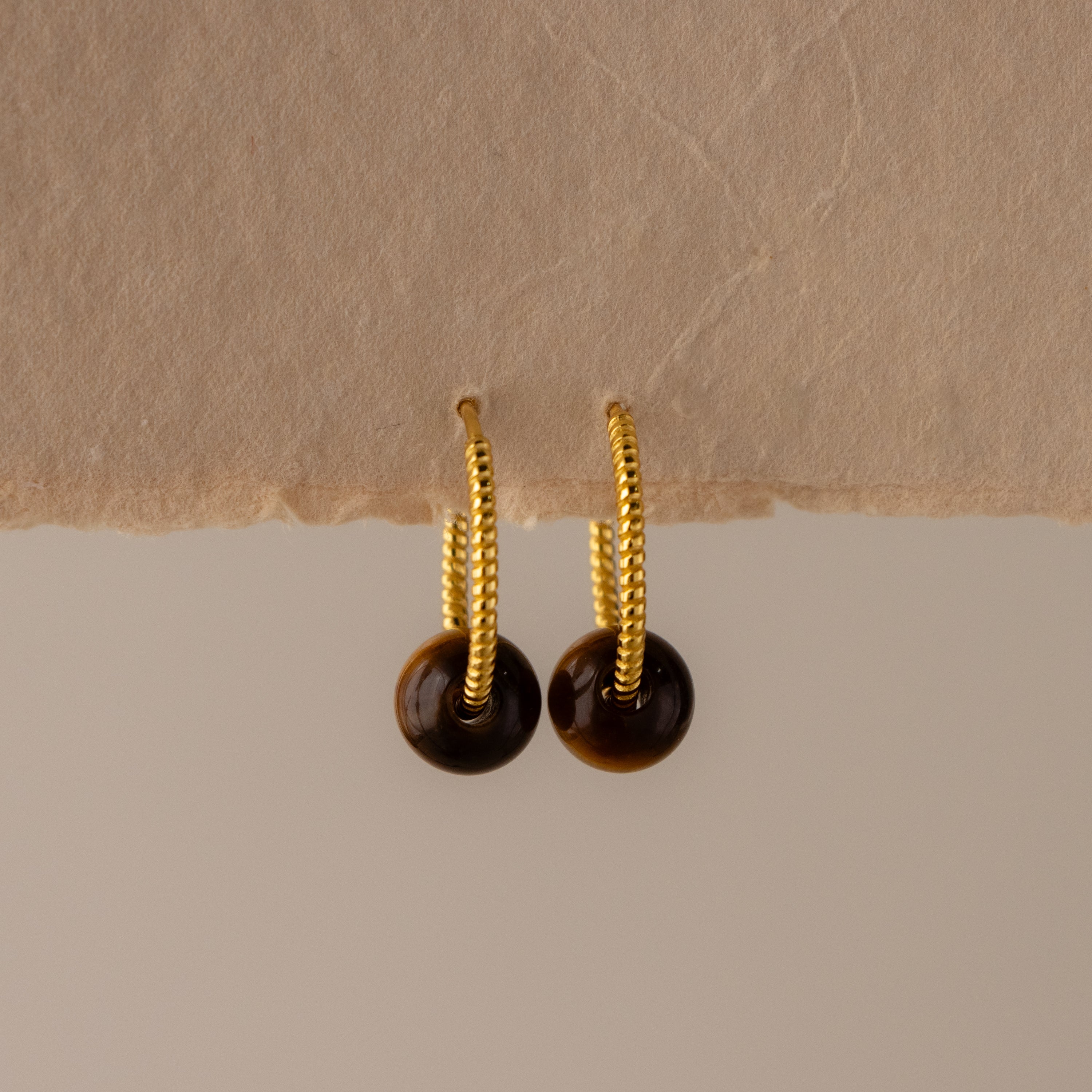 Twisted Tiger's Eye Bead Hoops feature gold textured hoops adorned with small round brown tiger's eye charms, displayed on a neutral textured surface.
