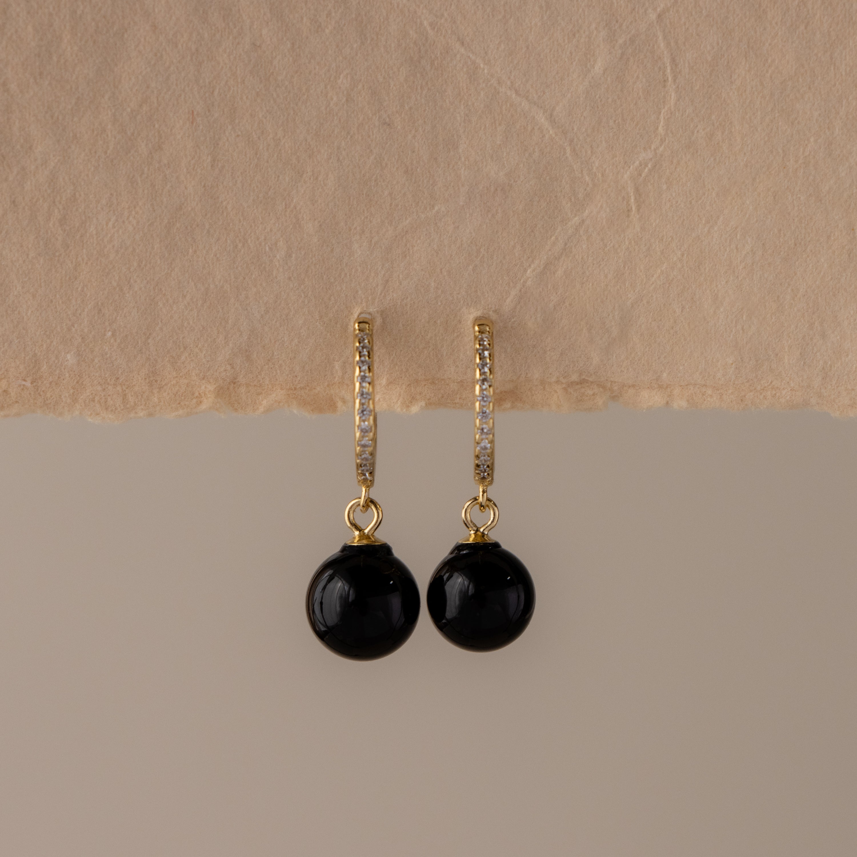 Pave Onyx Bead Drop Huggies, featuring gold hoops with black gemstone drops, displayed hanging on textured beige paper.