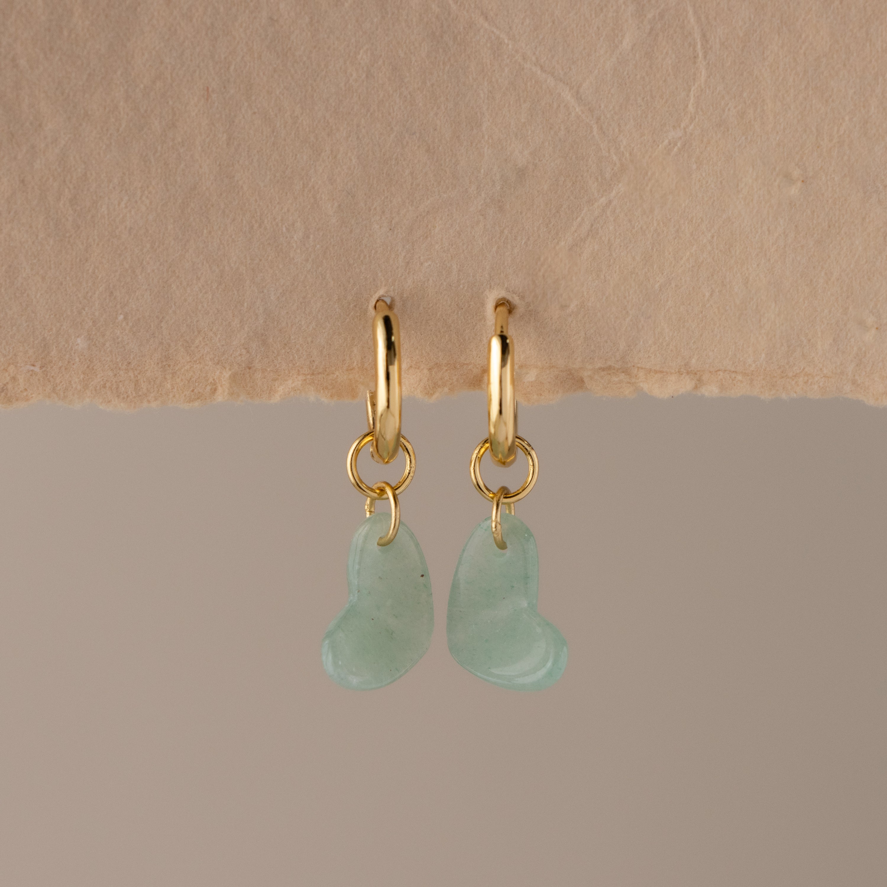 Aventurine Heart Drop Huggies: gold hoop earrings featuring light green, heart-shaped aventurine charms, elegantly displayed on a textured beige surface.