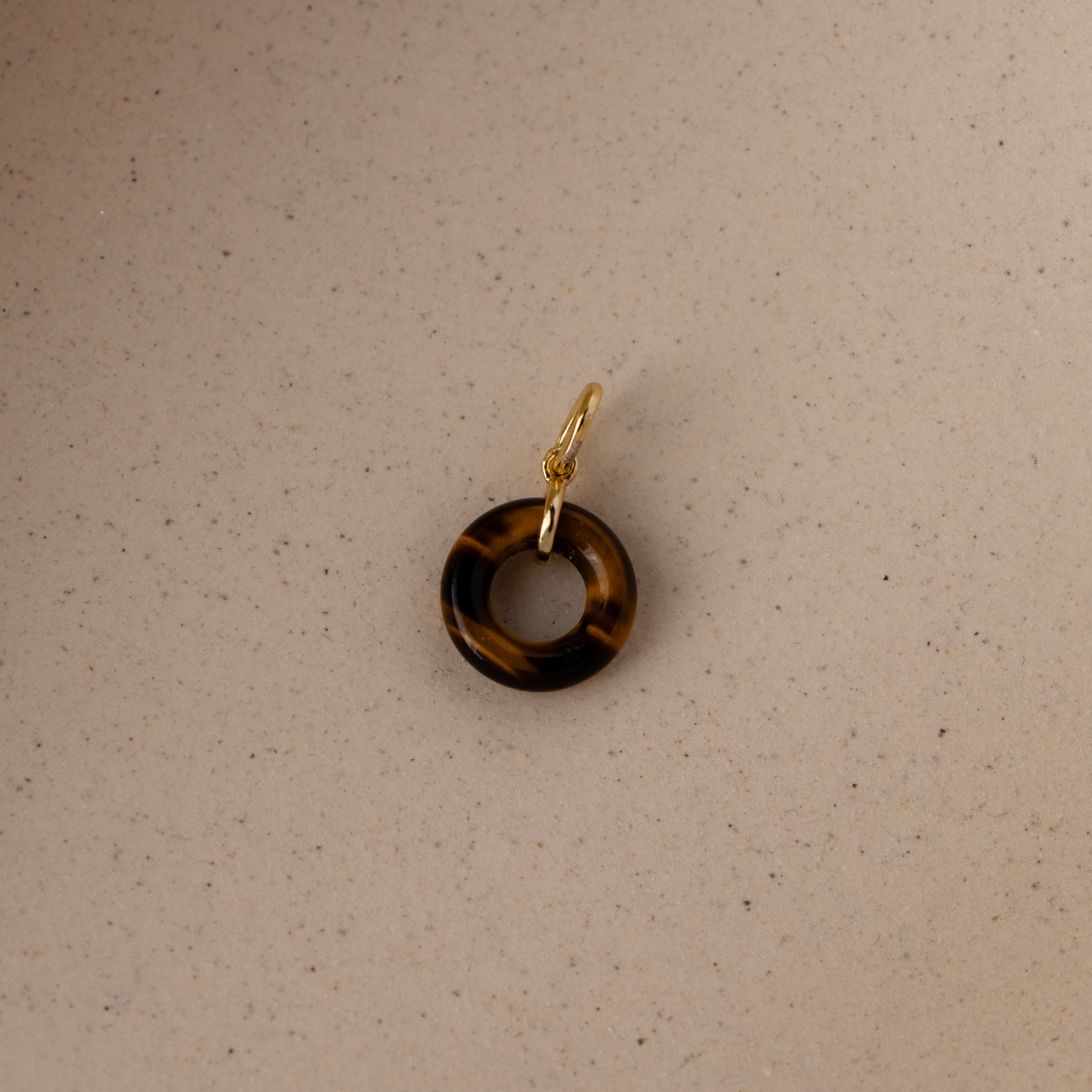 The Tiger's Eye Halo Charm is a brown tortoiseshell ring-shaped pendant with a gold jump ring, displayed on a beige textured surface.