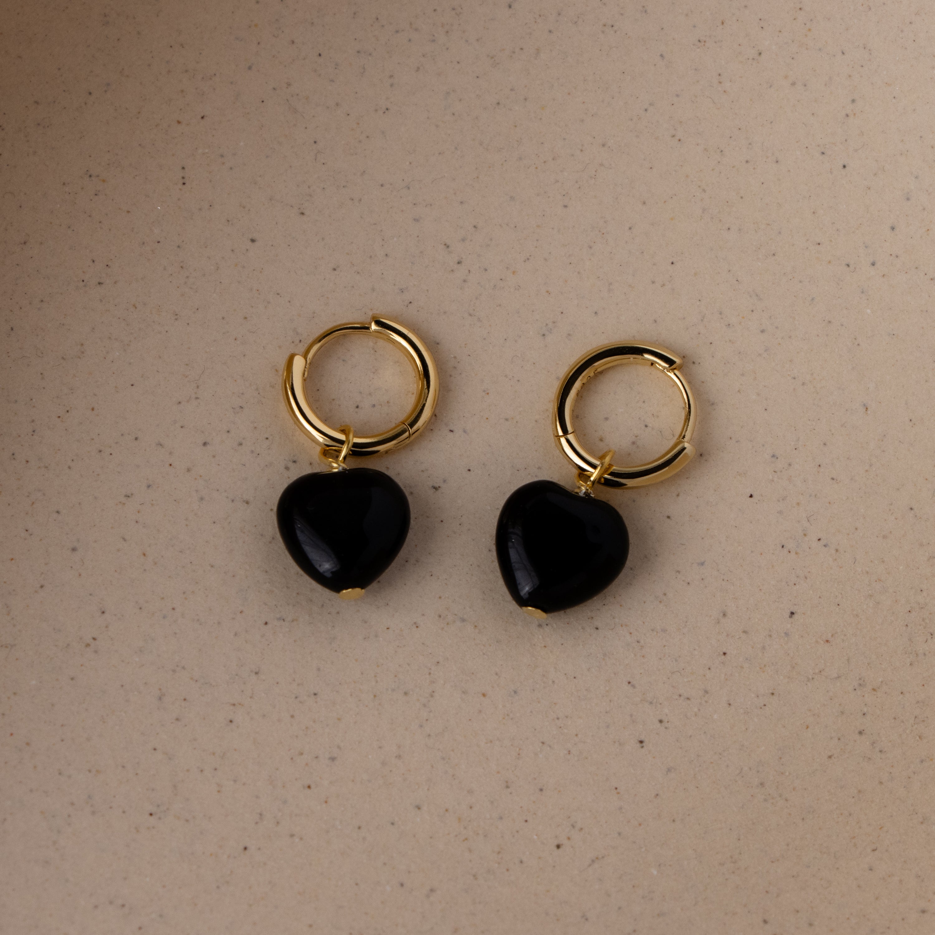 Onyx Heart Huggies: gold hoop earrings with black heart-shaped pendants displayed on a light beige surface.