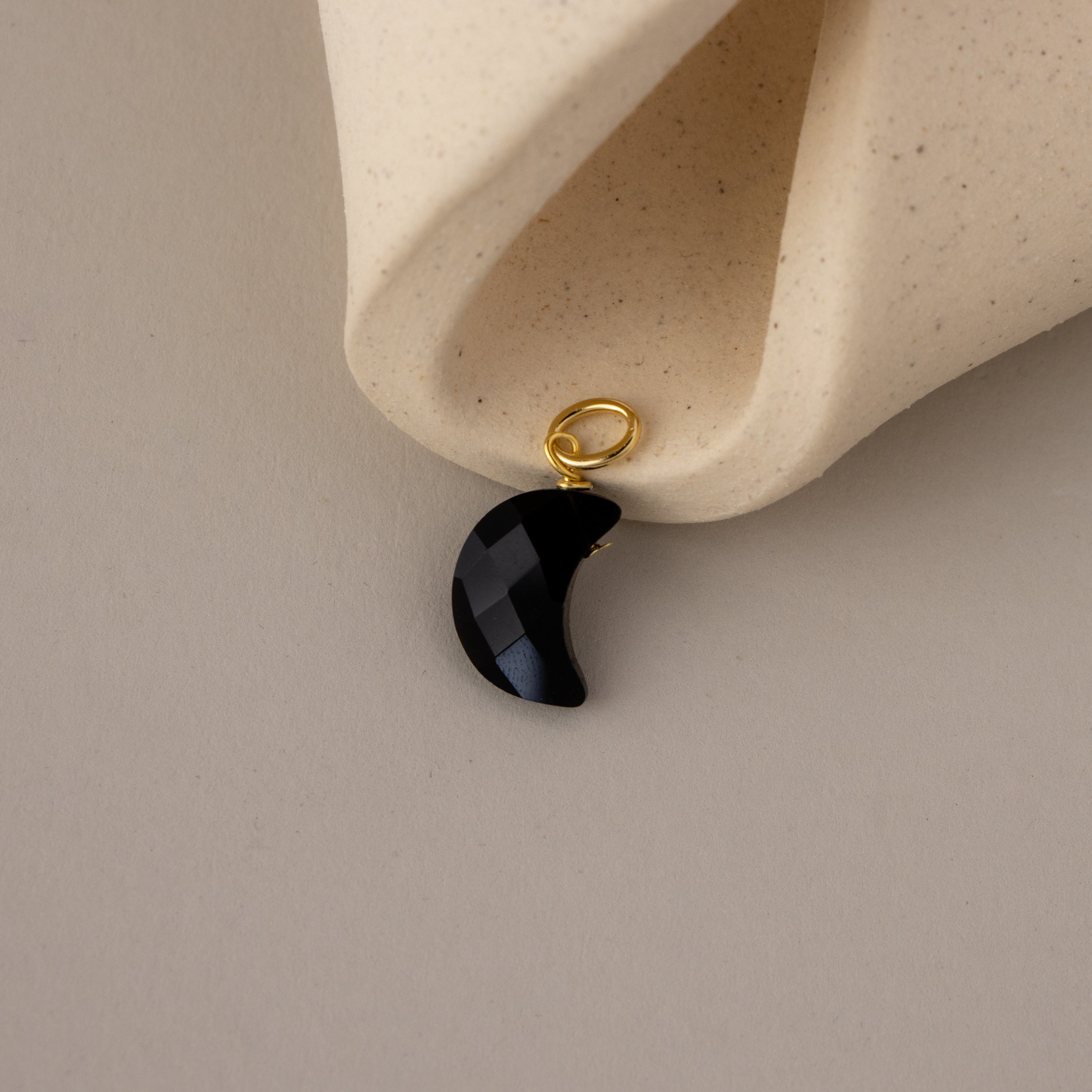 The Onyx Moon Charm, a black crescent pendant with a gold loop, is shown on a beige textured background.