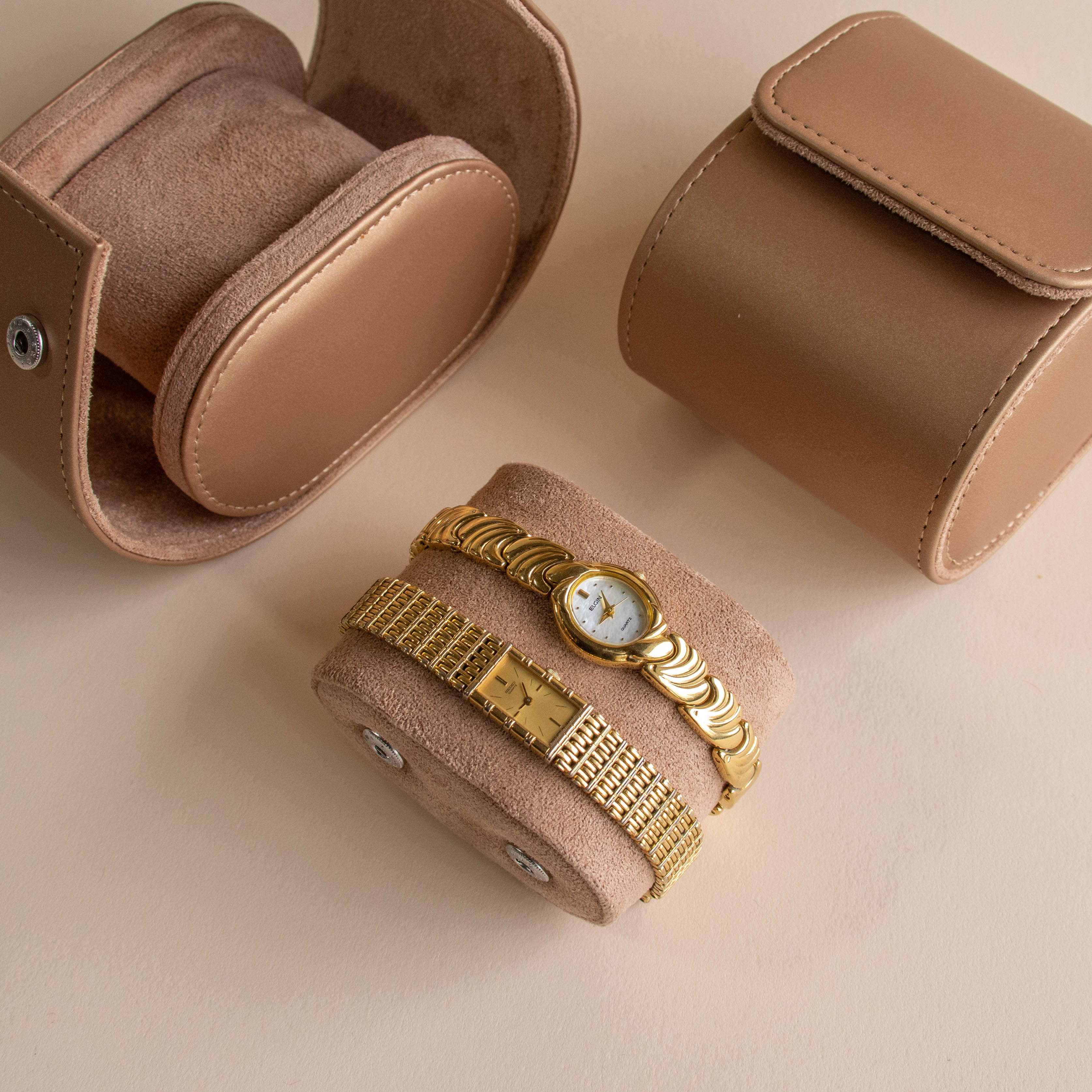 Two gold Pave Heart Timepiece watches, adorned with heart-shaped gemstones, are displayed on suede beside two brown suede cases against a beige background—capturing a sense of refined luxury.