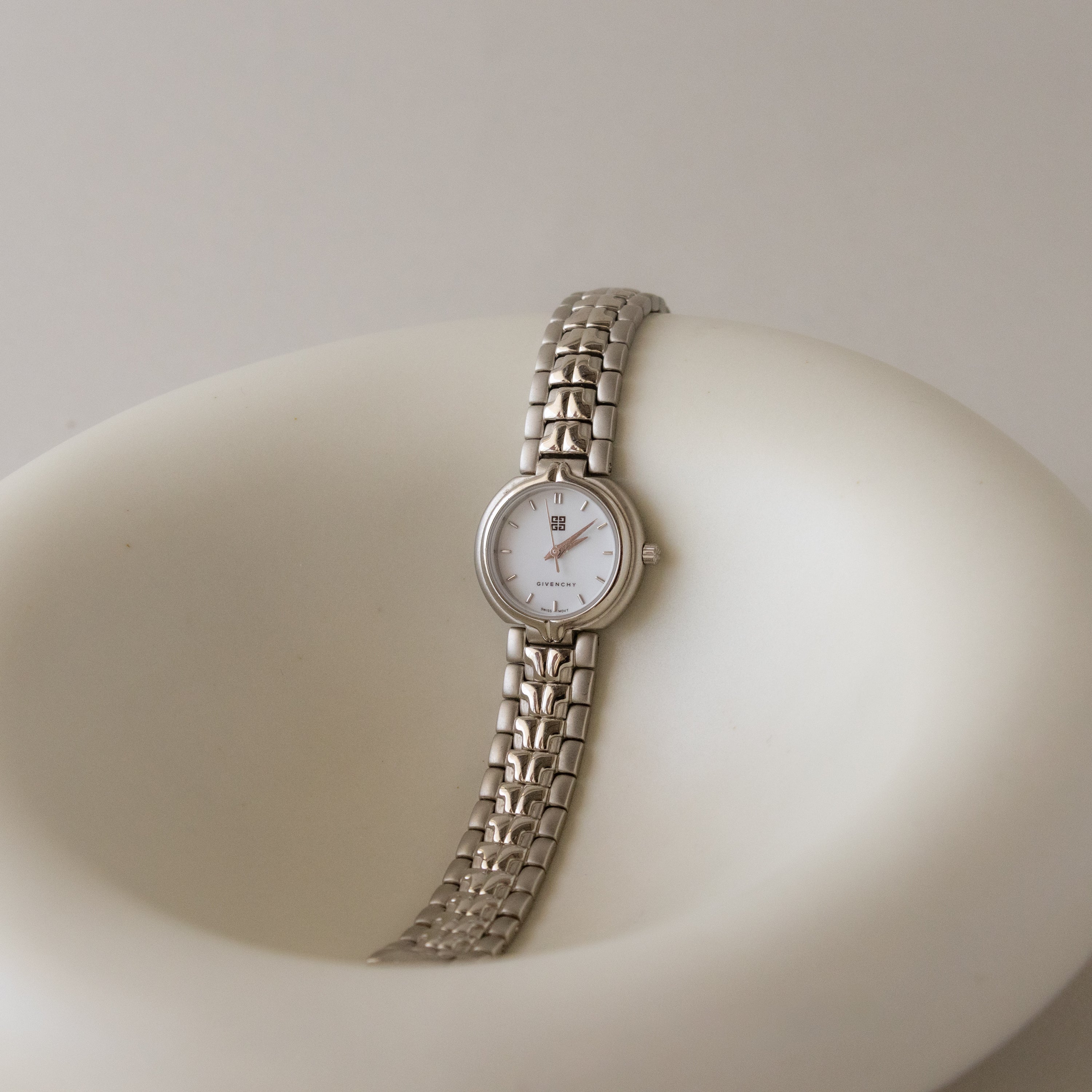 The Vintage Givenchy Swiss Bold Silver Watch, featuring a sleek silver case and white dial, is showcased on a smooth, curved white surface—an elegant statement piece for any collection.