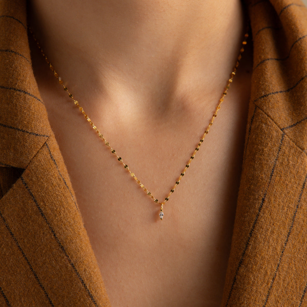 The Dainty Marquise Pendant Necklace, featuring a gold beaded chain and small pendant, is styled over a brown pinstripe blazer on a persons neck.