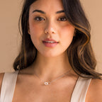 A woman with long brown hair wears a light dress and the Interlocking Pearl Necklace, posing against a beige background.