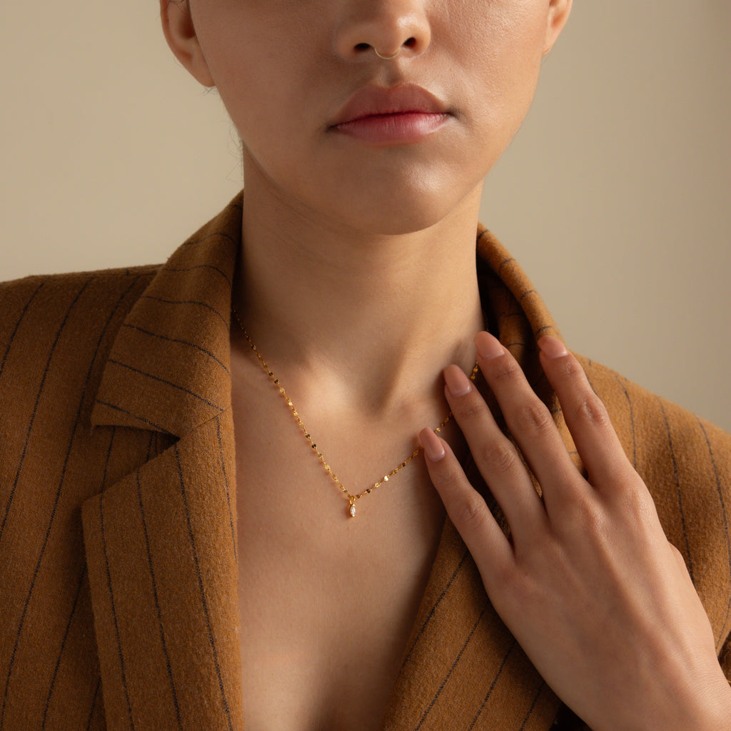A woman in a brown pinstripe blazer touches the Dainty Marquise Pendant Necklace, featuring a small gold marquise pendant on an elegant mirror twist chain.