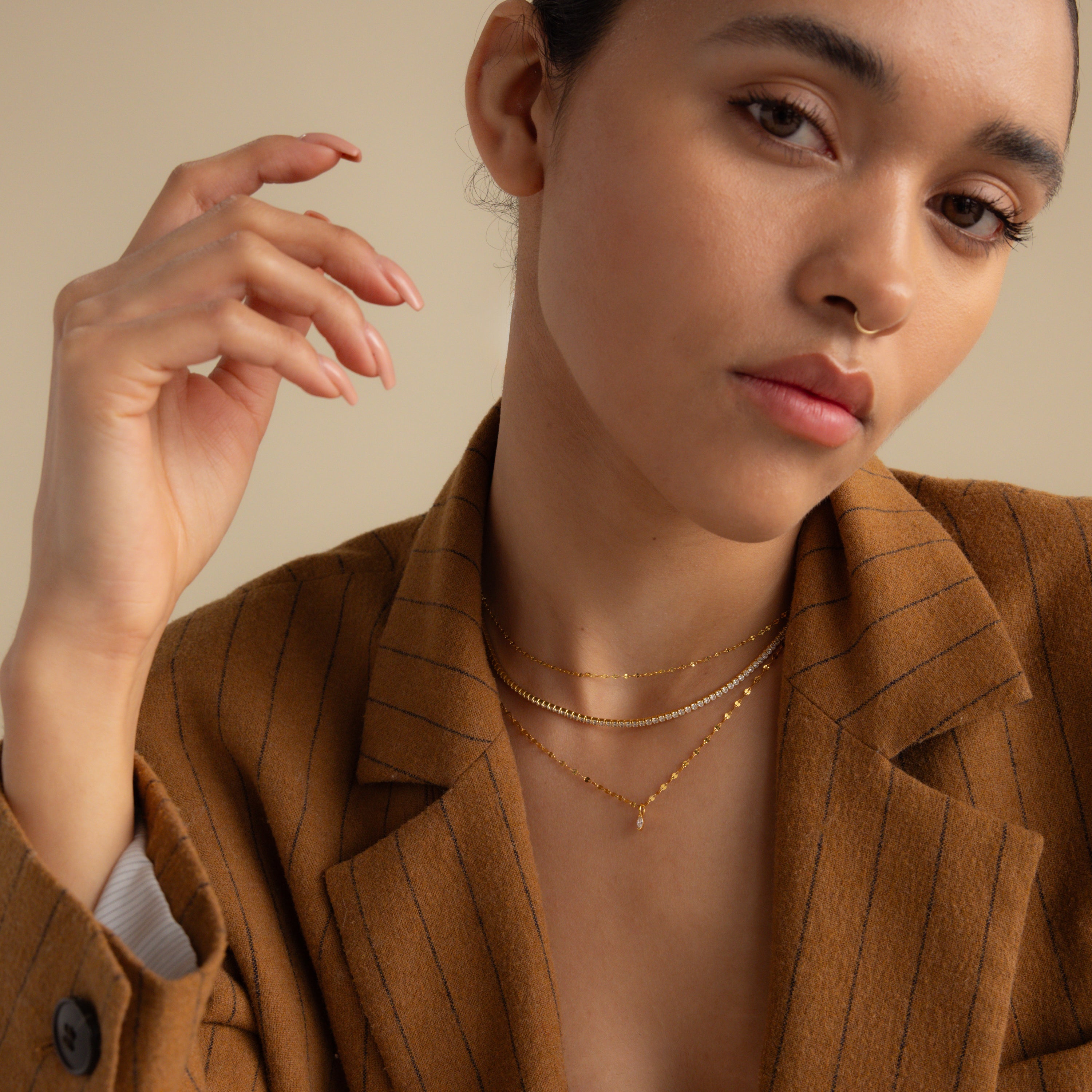 Woman in a brown pinstripe blazer wearing layered gold necklaces, including the Dainty Marquise Pendant Necklace in 18K Gold, posing with her hand raised near her face.