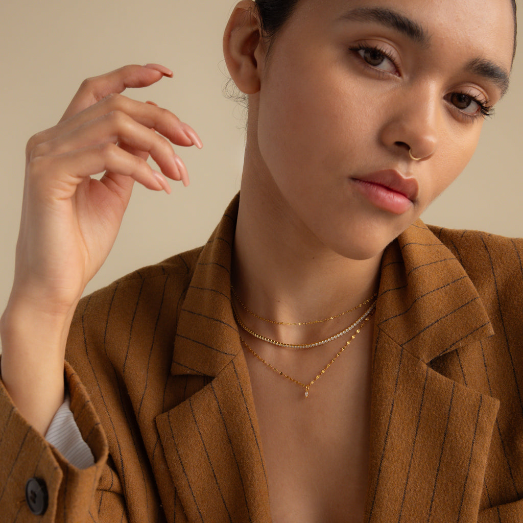 Woman in a brown pinstripe blazer, wearing layered gold necklaces including the Dainty Marquise Pendant Necklace, poses with her hand near her face.