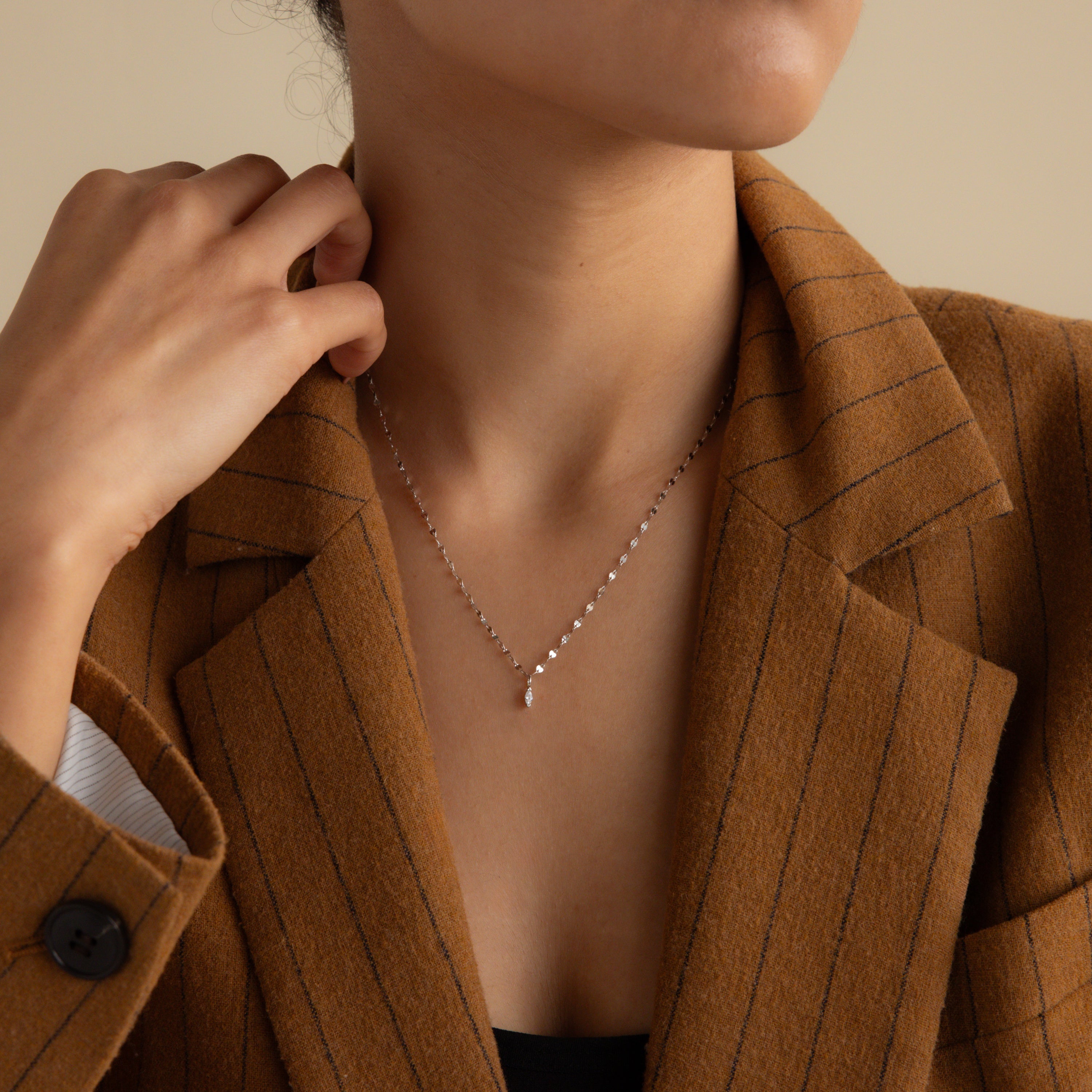 A woman in a brown pinstripe blazer wears the Dainty Marquise Pendant Necklace in Silver with a mirror twist chain, her hand resting gently near her neck.