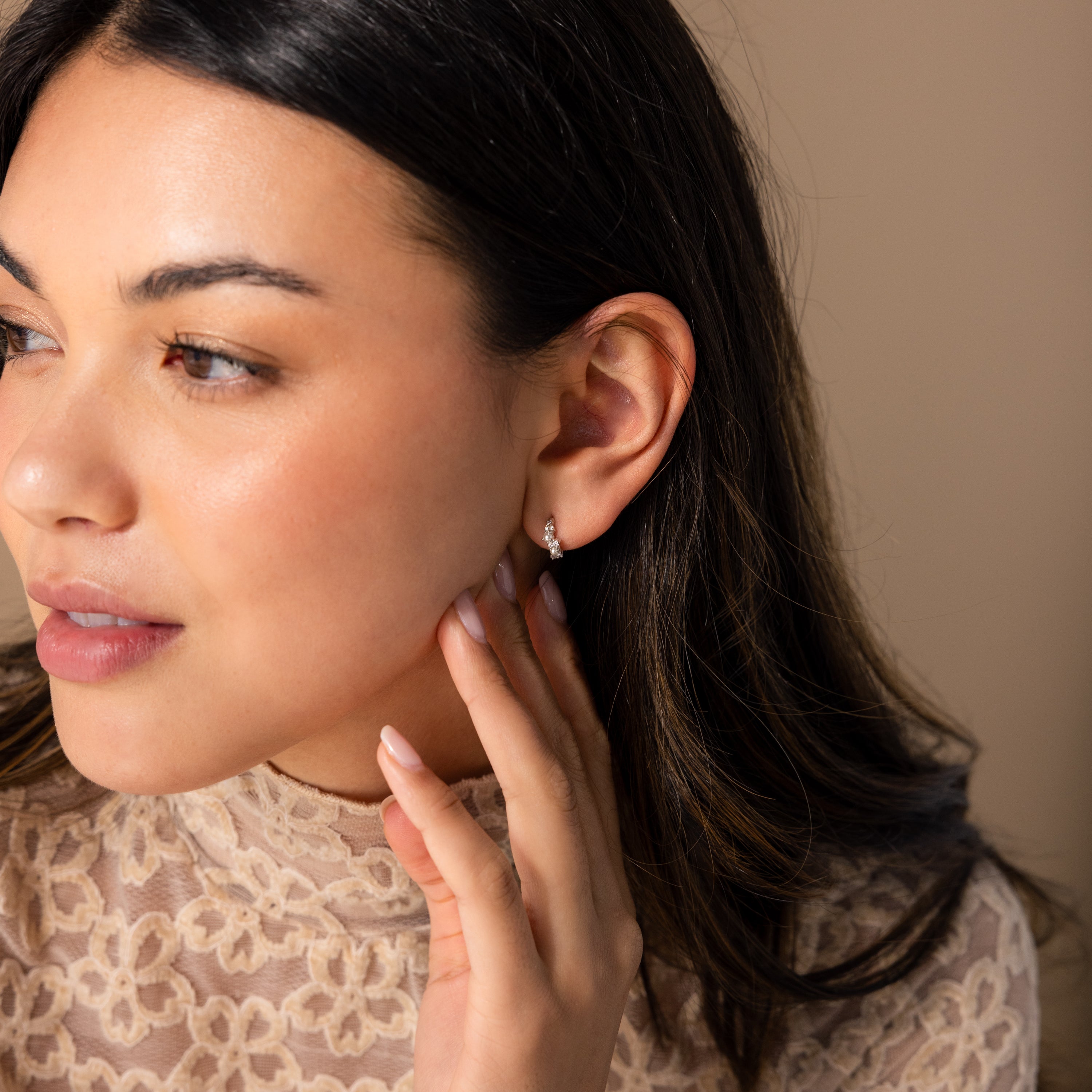 A woman with long dark hair, wearing a lace top, touches her Pearl Diamond Cluster Huggies in Sterling Silver while gazing to the side.