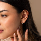 A woman touches her neck, highlighting Pearl Diamond Cluster Huggies in Sterling Silver worn in her pierced ear against a neutral background.