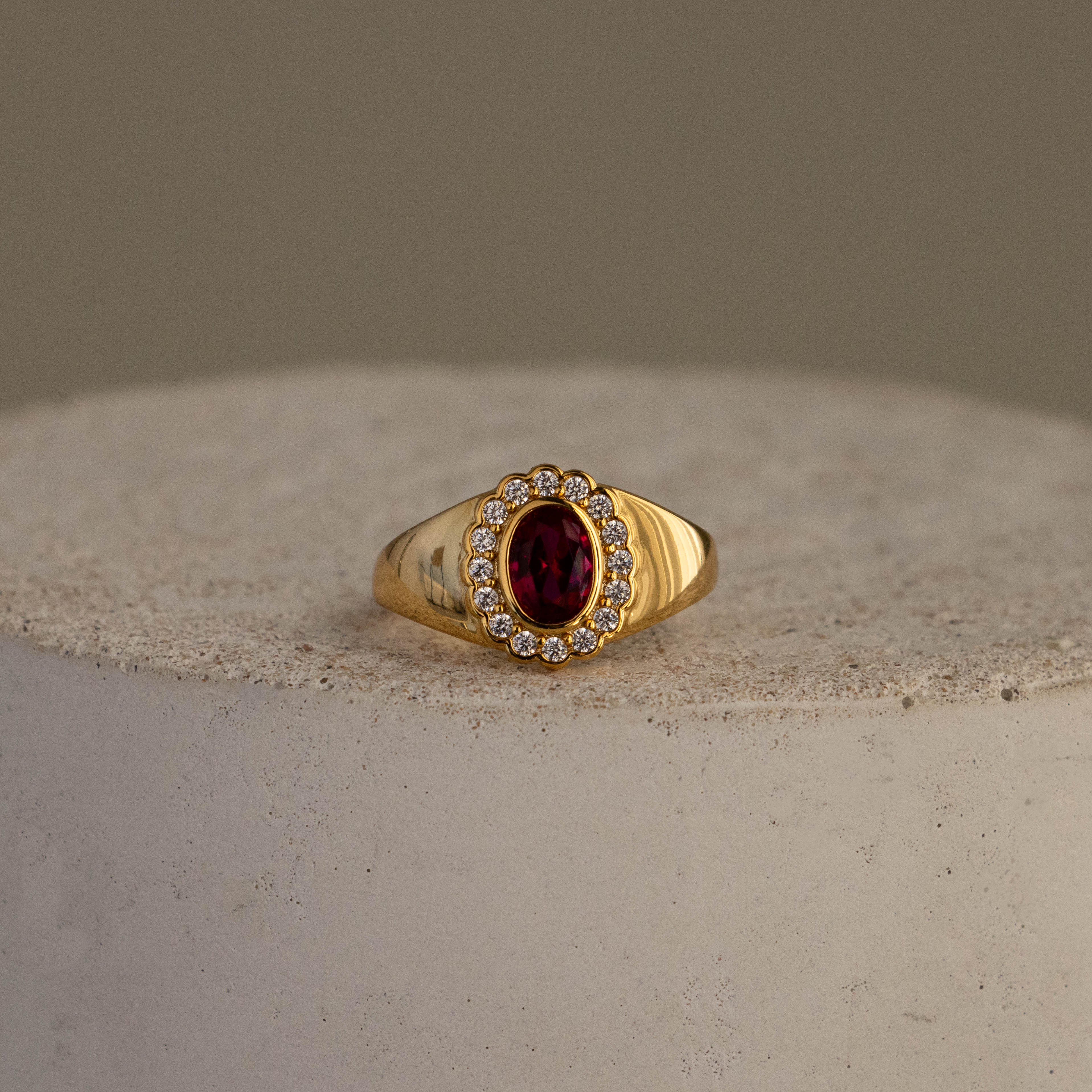Diamond  Ruby Heritage Signet Ring featuring an oval red gemstone encircled by sparkling diamonds, shown on a textured surface.