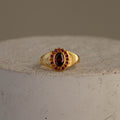 The Garnet  Ruby Heritage Signet Ring features an oval red gemstone encircled by smaller rubies, showcased on a textured surface.