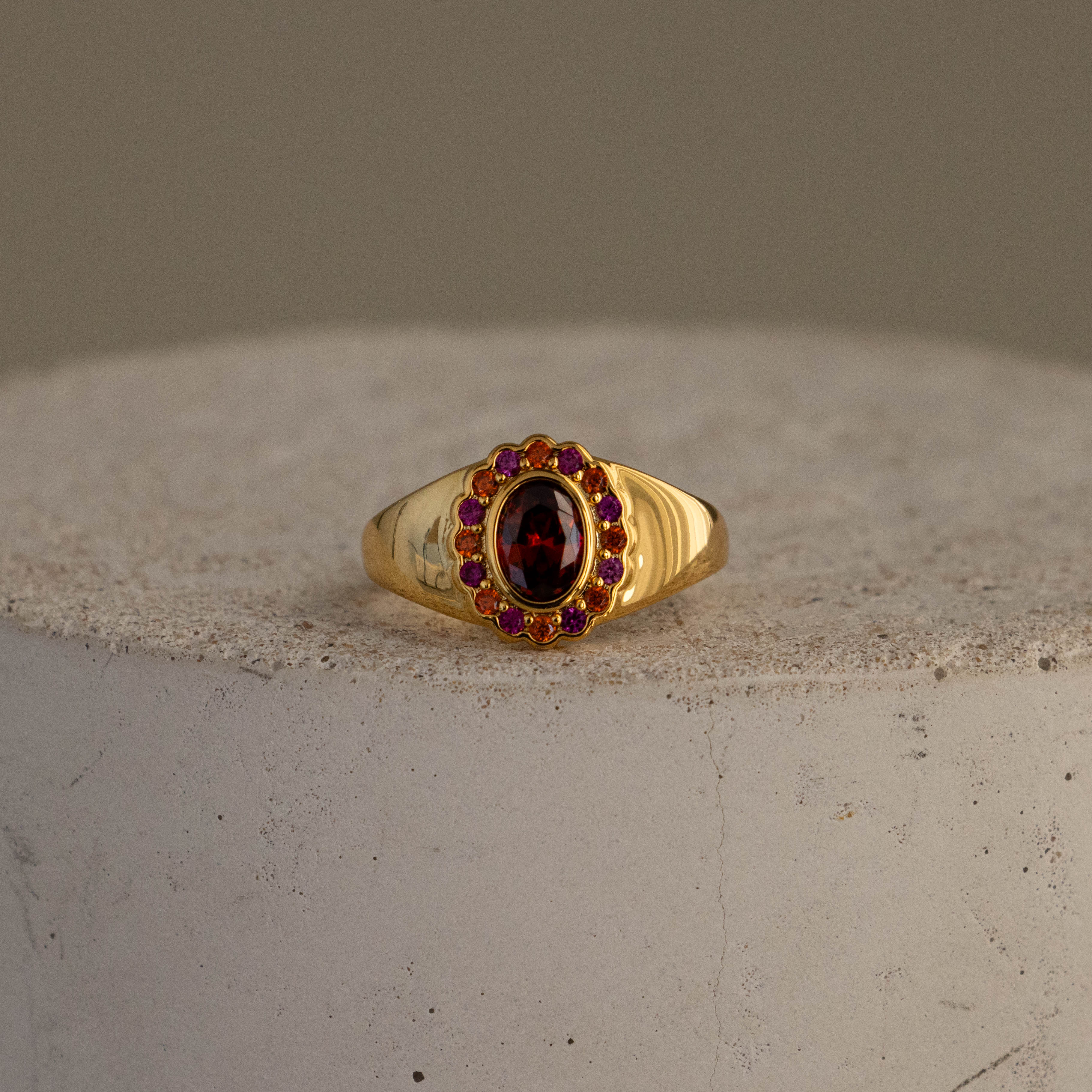 The Garnet  Ruby Heritage Signet Ring features an oval red gemstone encircled by smaller rubies, showcased on a textured surface.